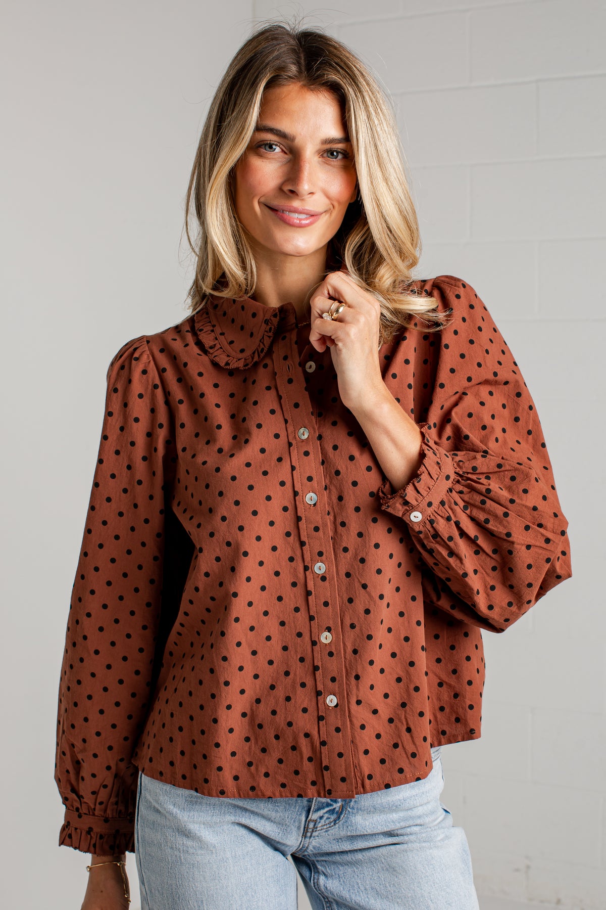 A woman wearing a rust-brown long-sleeve blouse with black polka dots, a Peter Pan collar, and voluminous ruffled sleeves, paired with light-wash blue jeans against a white brick wall.