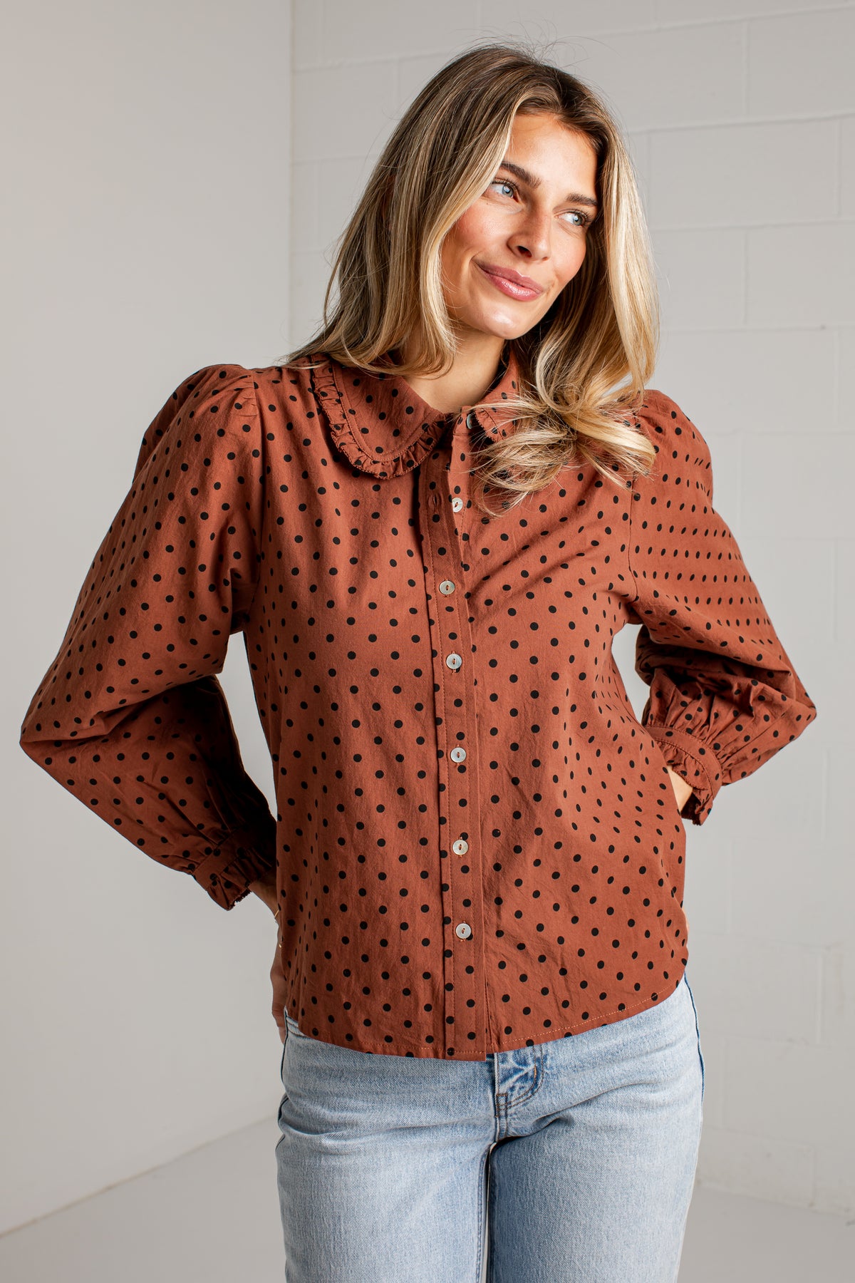 A woman wearing a rust-brown long-sleeve blouse with black polka dots and a large Peter Pan collar, paired with light-wash blue jeans, standing against a white brick wall.