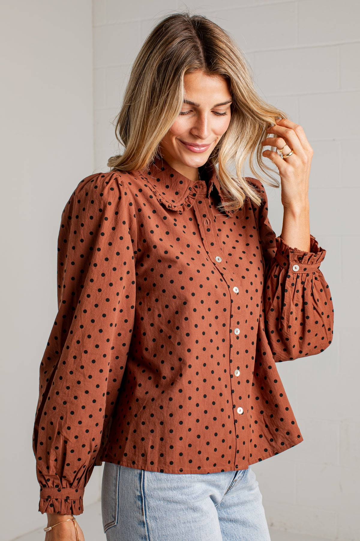A woman wearing a rust-brown long-sleeve blouse with black polka dots, featuring a ruffled collar and voluminous puff sleeves, paired with light blue jeans.