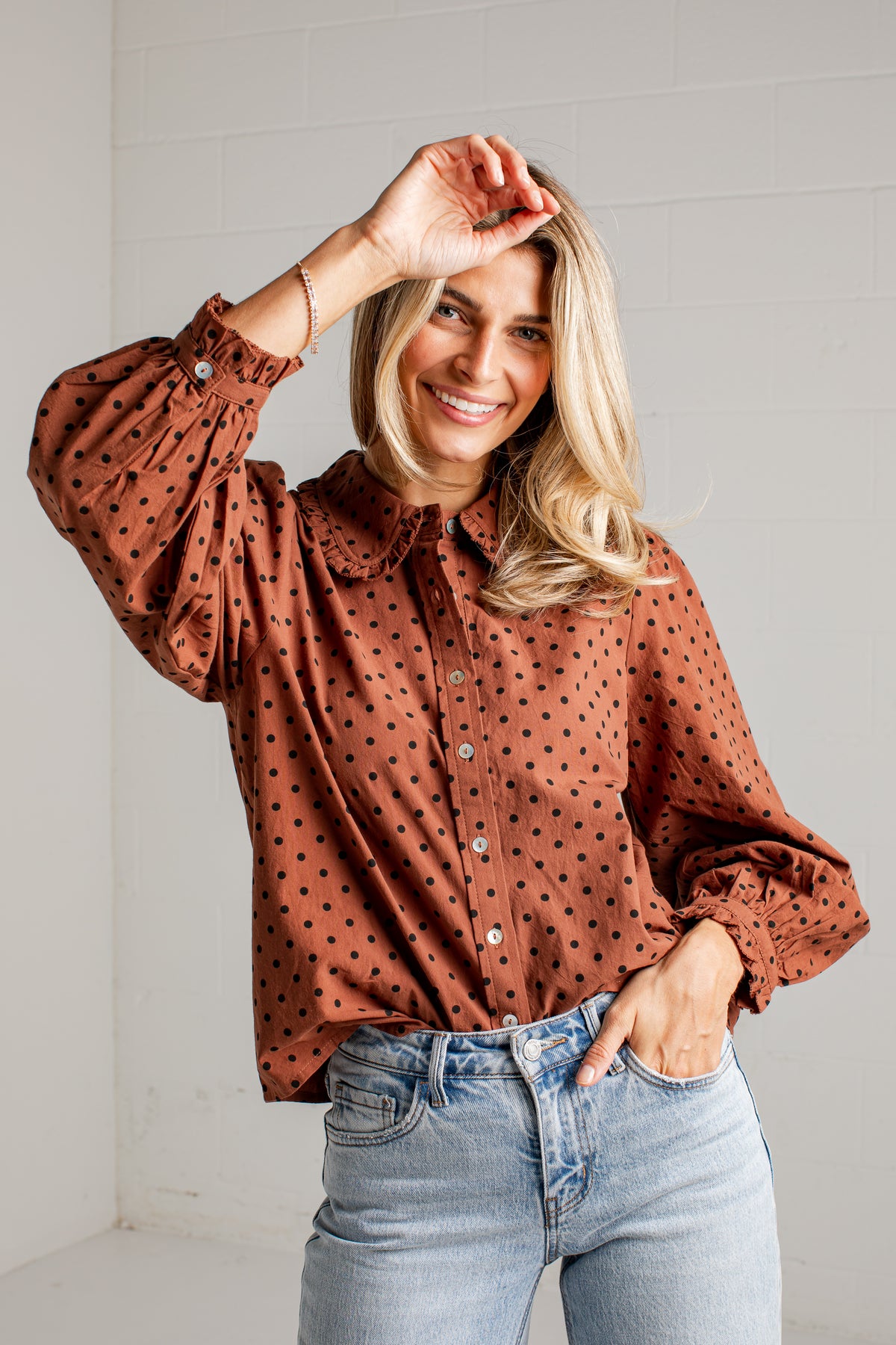 A smiling blonde woman wearing a rust-brown long-sleeve button-up blouse with black polka dots and light-wash blue jeans, posing in a bright indoor setting.