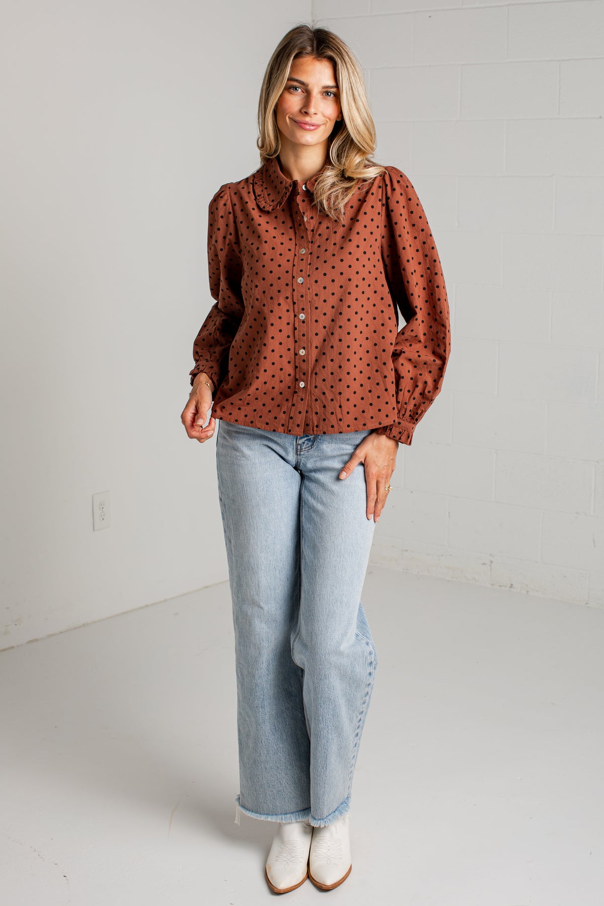 A woman modeling a rust-brown long-sleeve button-up blouse with a black polka dot pattern, paired with light-wash wide-leg jeans and white ankle boots, standing against a white brick wall.