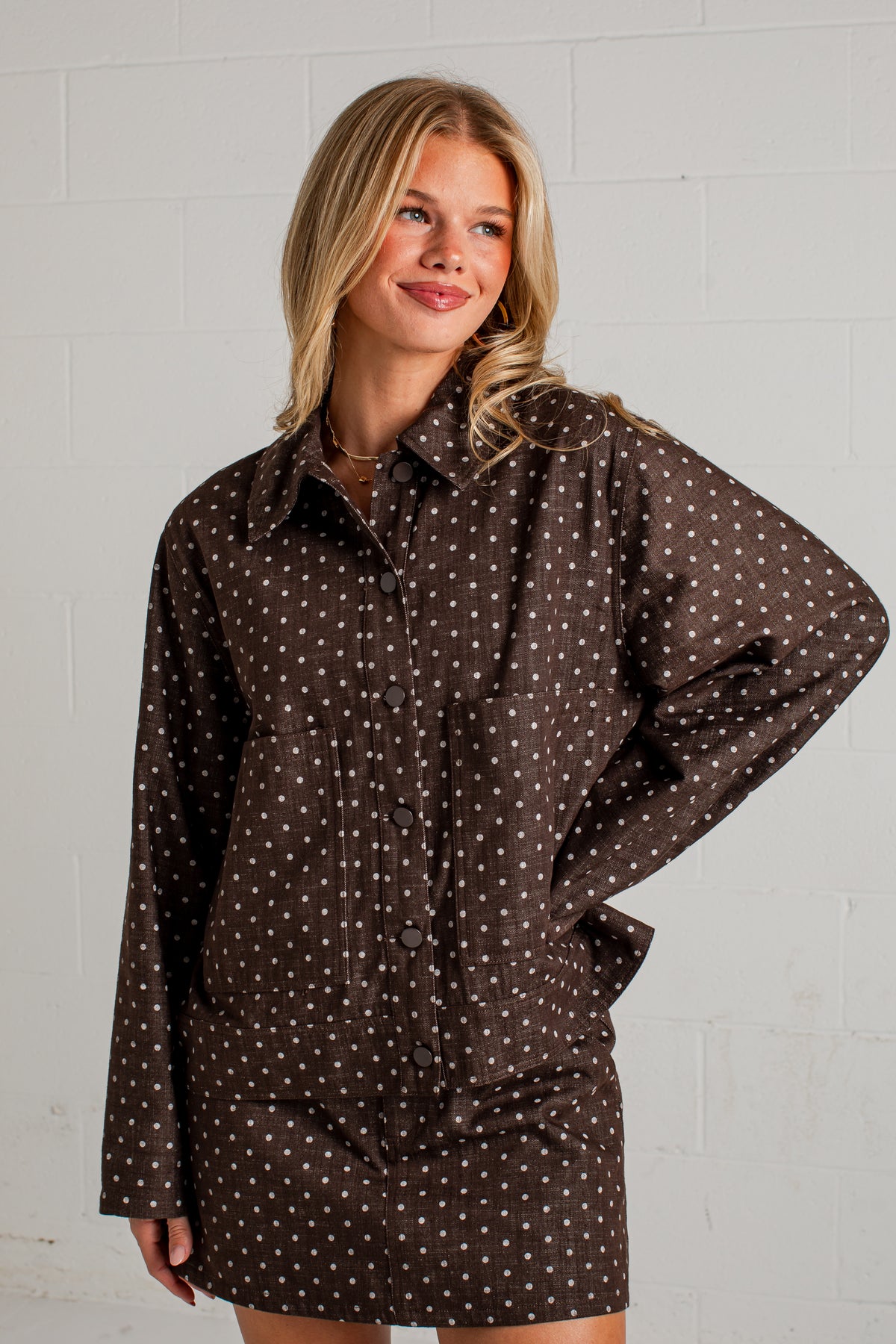 A woman modeling a dark brown long-sleeve button-up shirt dress with a white polka dot pattern, standing against a white brick wall.