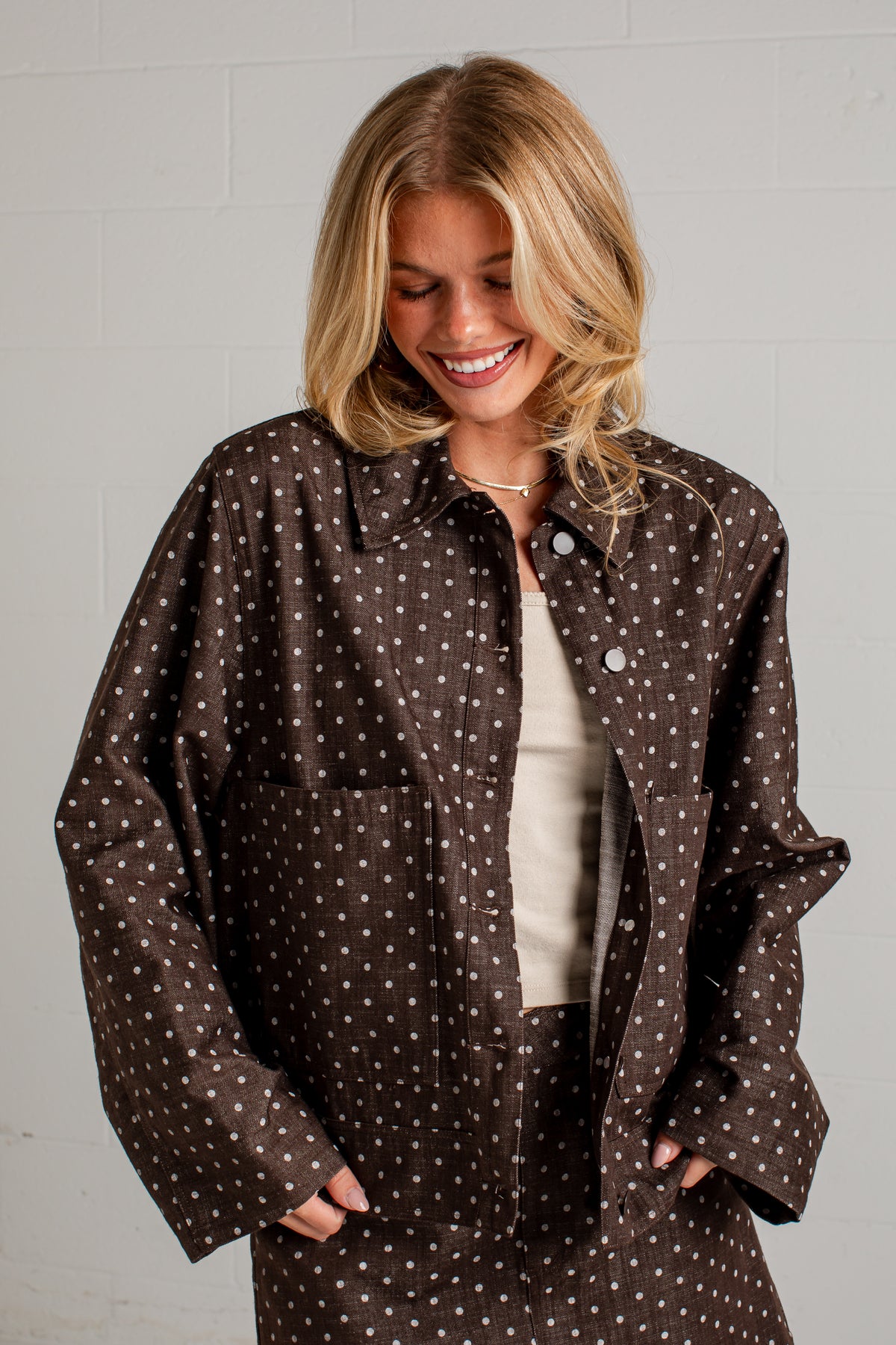 A woman wearing a brown button-up shirt jacket with a white polka dot pattern, layered over a cream top, standing against a white brick wall.