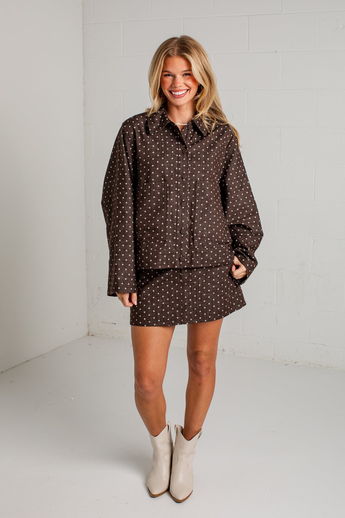 A woman modeling a matching brown and white polka dot shirt and skirt set, paired with beige ankle boots, standing against a white brick wall.