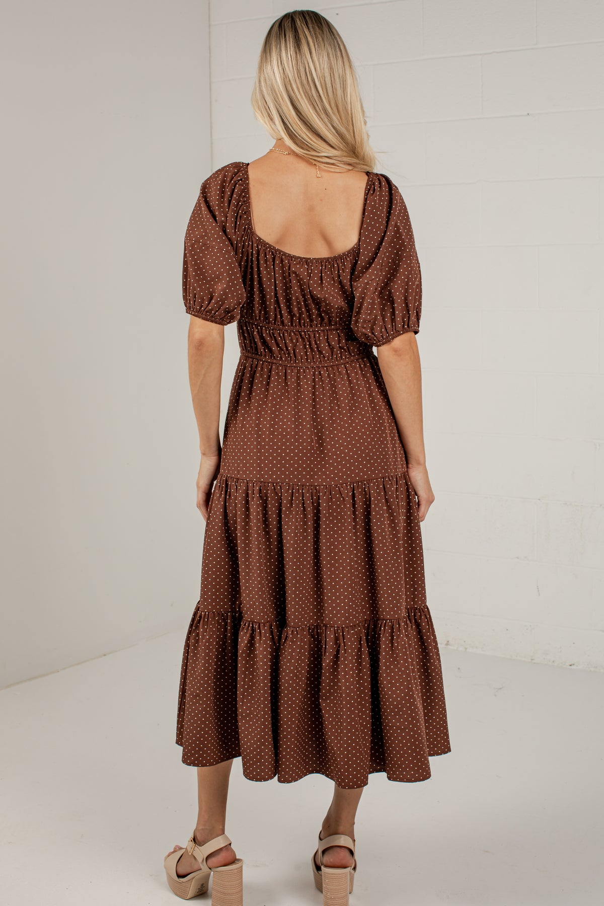 A woman viewed from behind wearing a brown tiered midi dress with a small white polka dot pattern, puff sleeves, and a scoop neckline, paired with beige platform sandals against a white brick wall background.