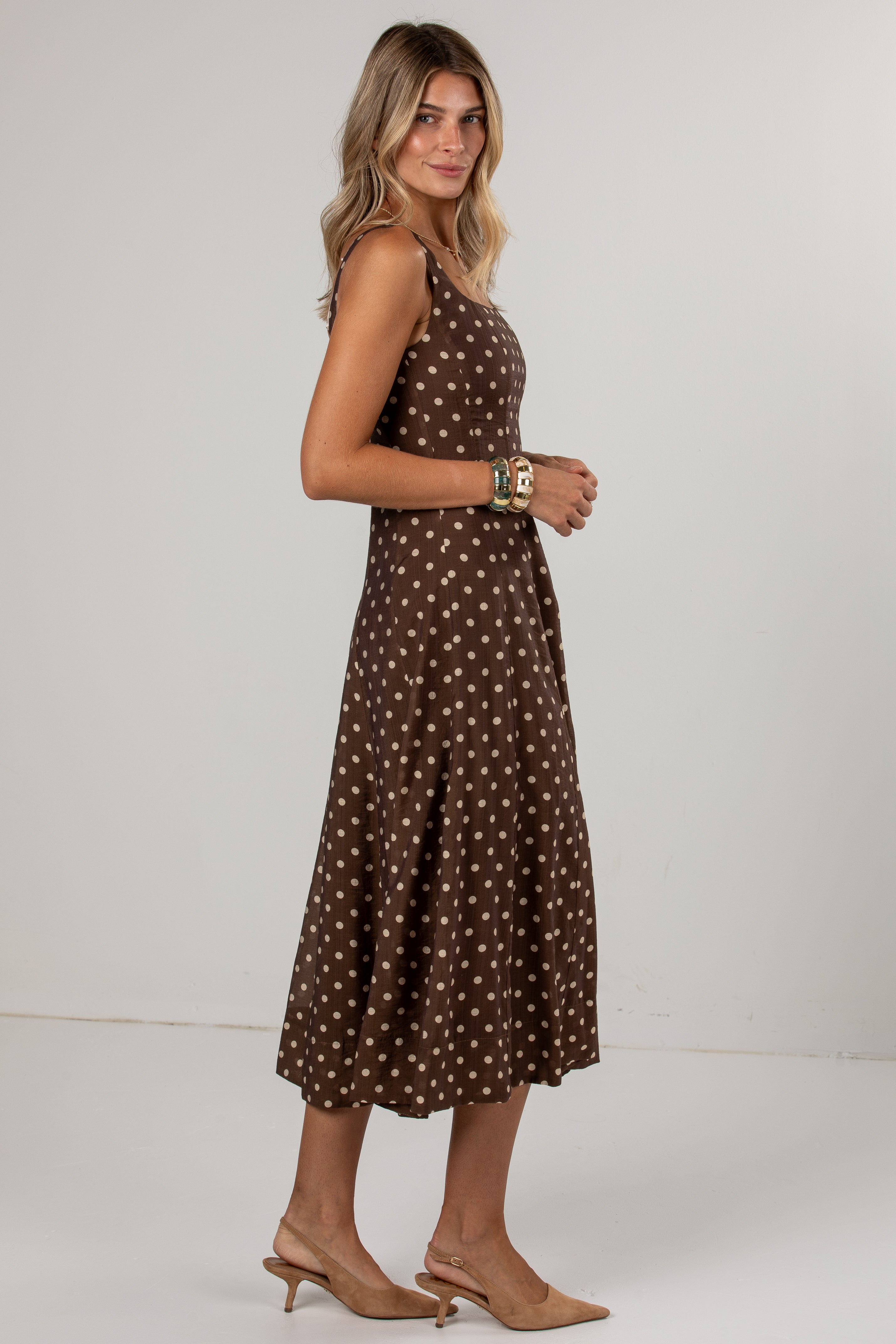 A woman modeling a sleeveless brown midi dress with a white polka dot pattern, paired with beige slingback heels, against a plain light gray studio background.