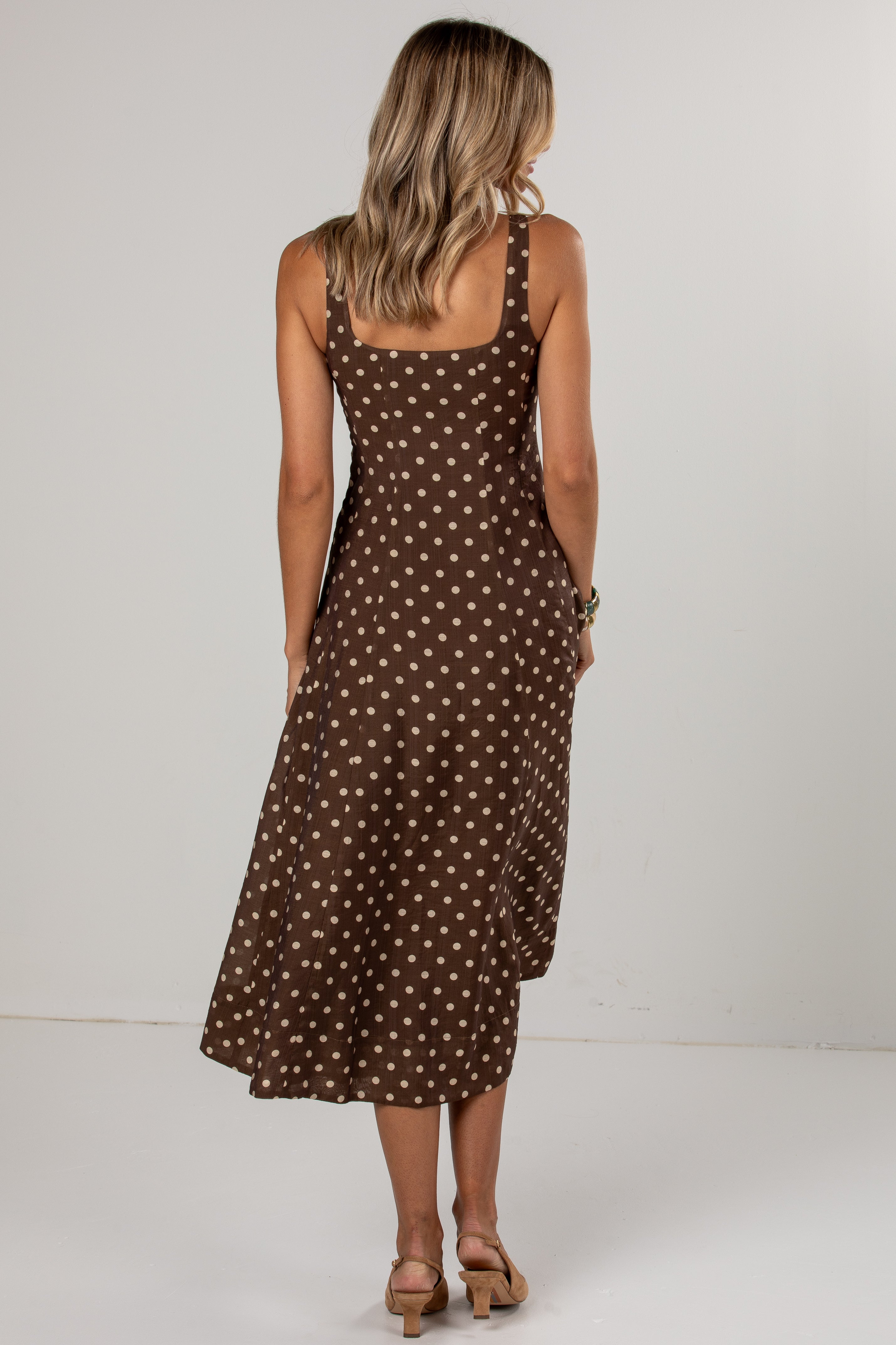 A woman viewed from behind wearing a brown midi dress with a white polka dot pattern, paired with tan heeled sandals, set against a plain light gray studio background.