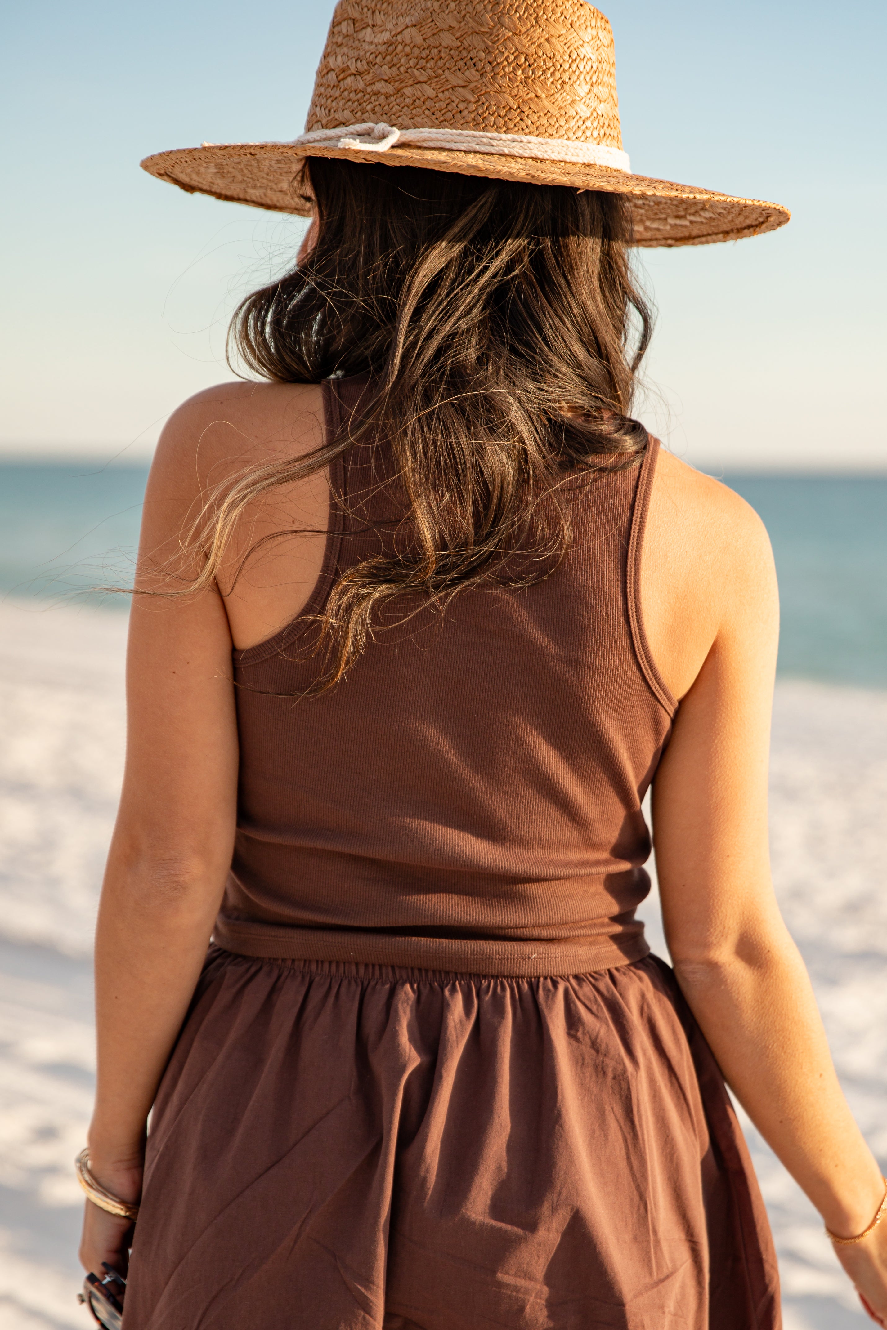 Woman in a brown tank top and brown skirt and hat standing on a beach