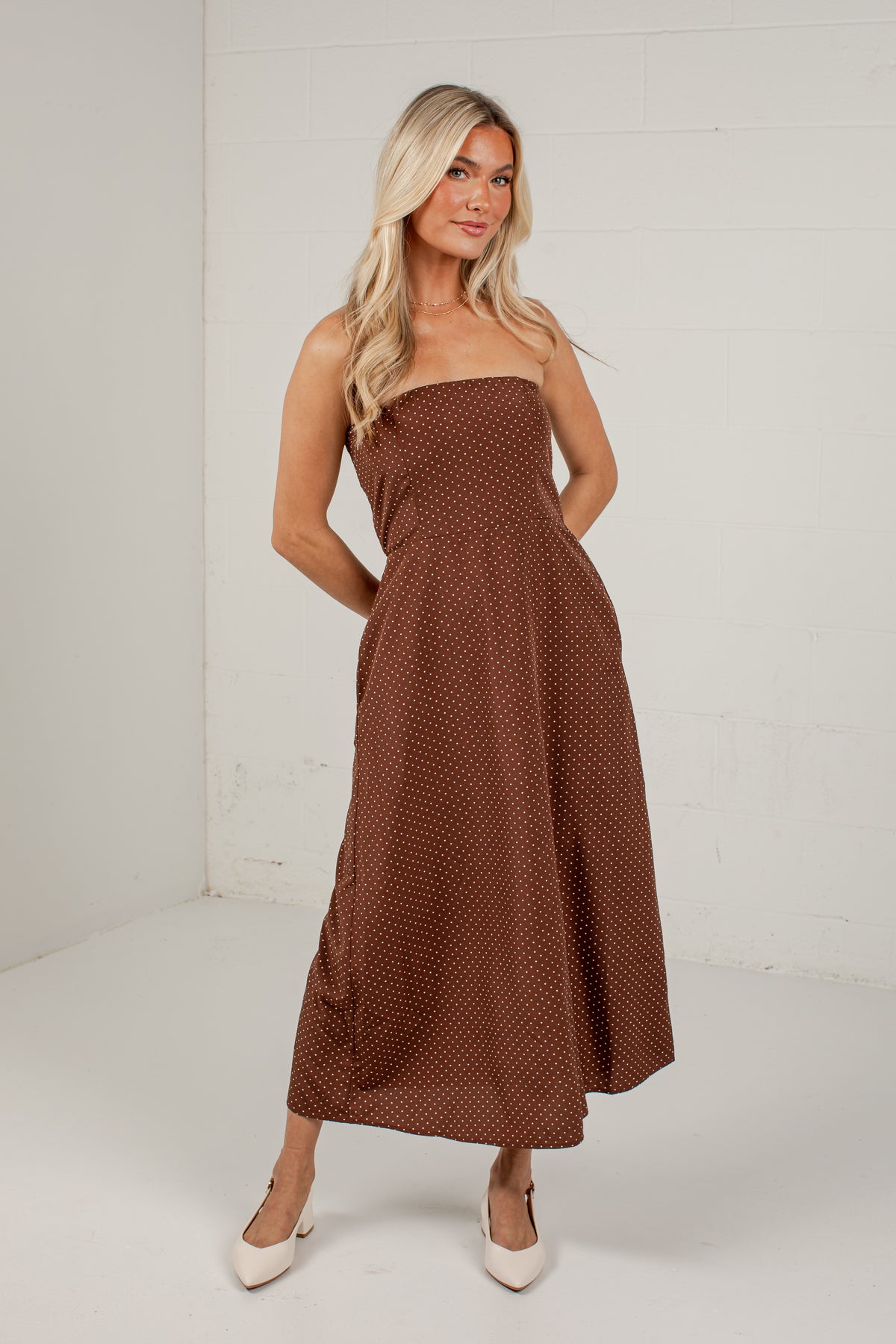 A woman modeling a strapless brown maxi dress with a small white polka dot pattern, standing against a white brick wall.