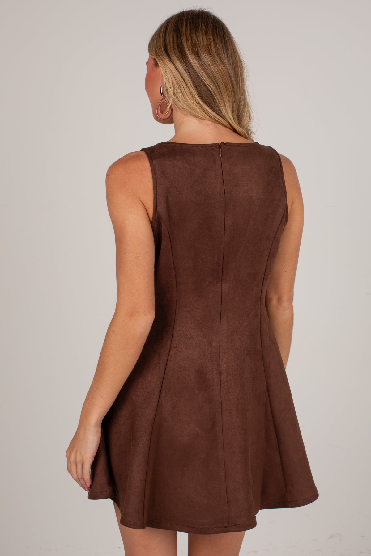A woman shown from the back wearing a sleeveless brown dress with a flared skirt, photographed against a plain light gray studio background.