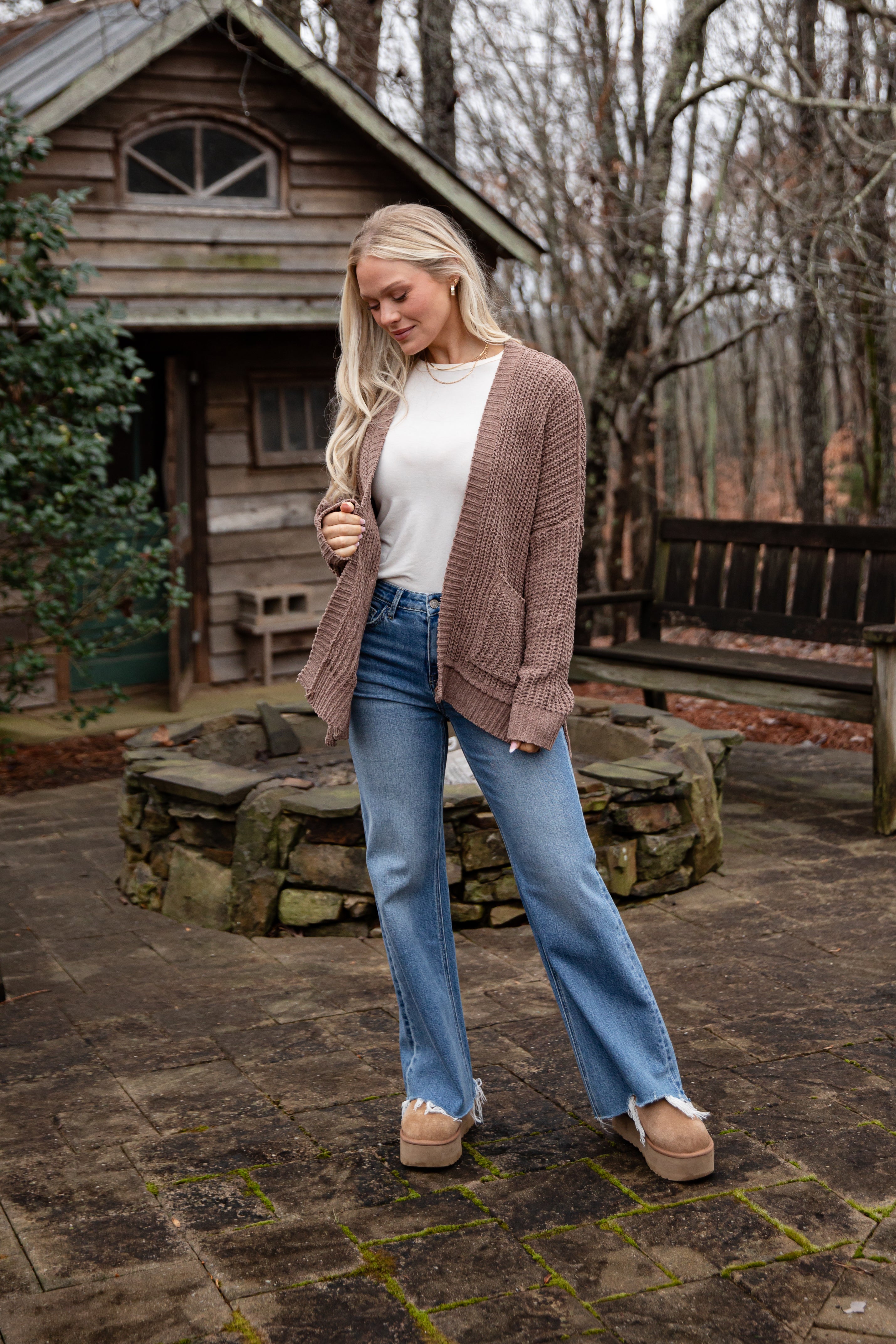 Woman wearing a brown cardigan, white shirt, blue jeans, and beige boots standing in front of a wooden cabin.