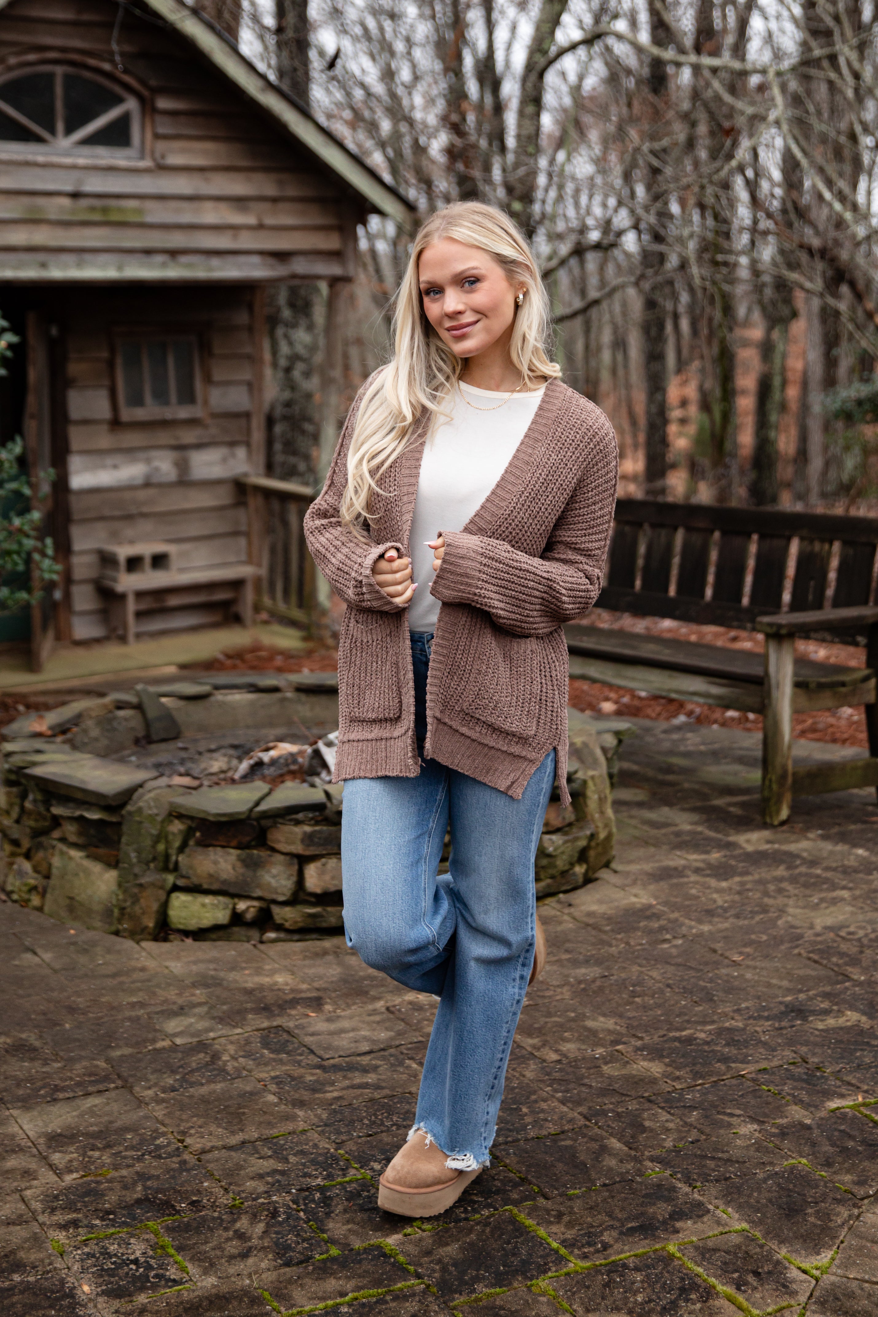 Woman standing outdoors in front of a wooden cabin, wearing a brown cardigan and blue jeans.