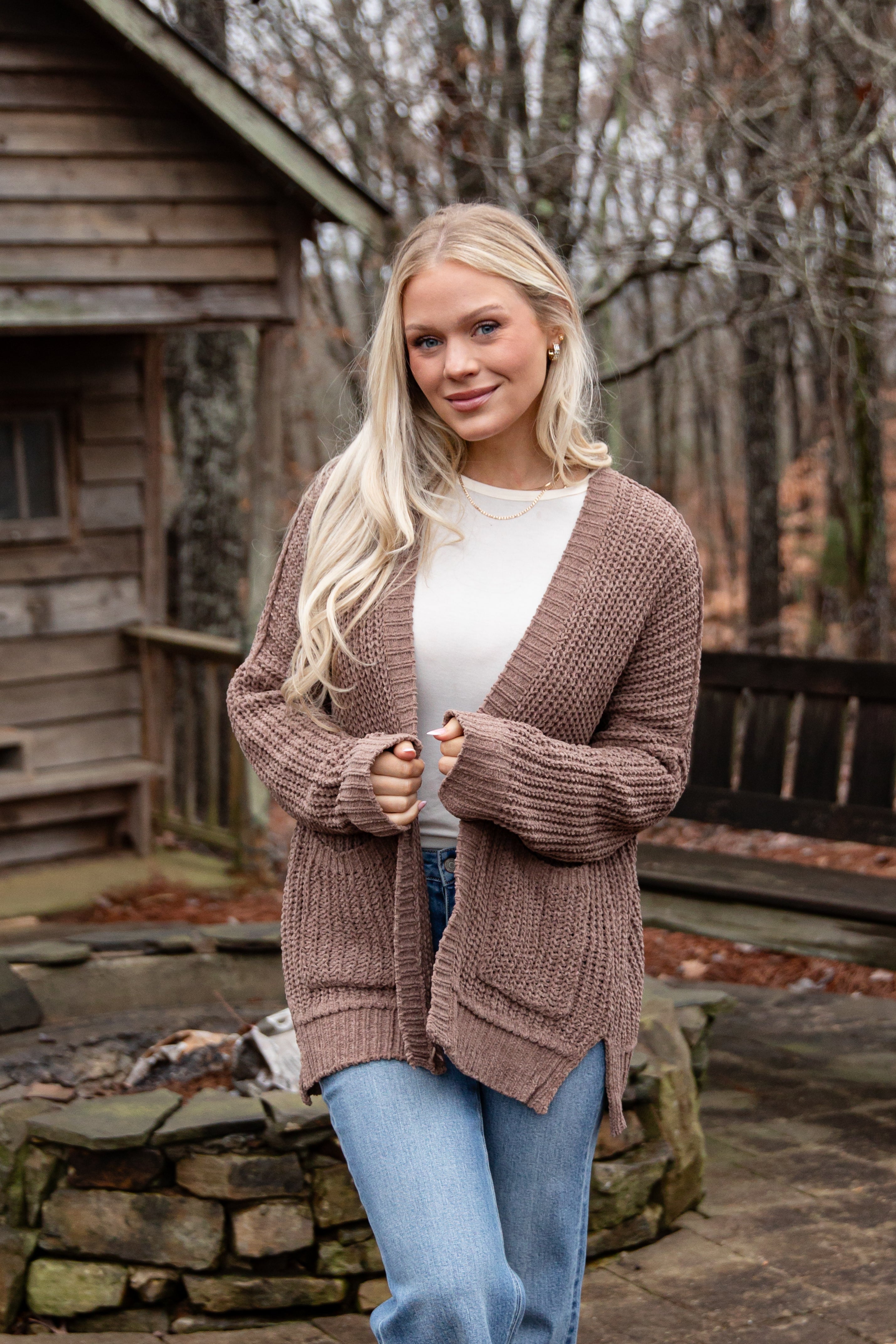 Woman wearing a brown cardigan over a white shirt and blue jeans, standing in front of a wooden cabin.