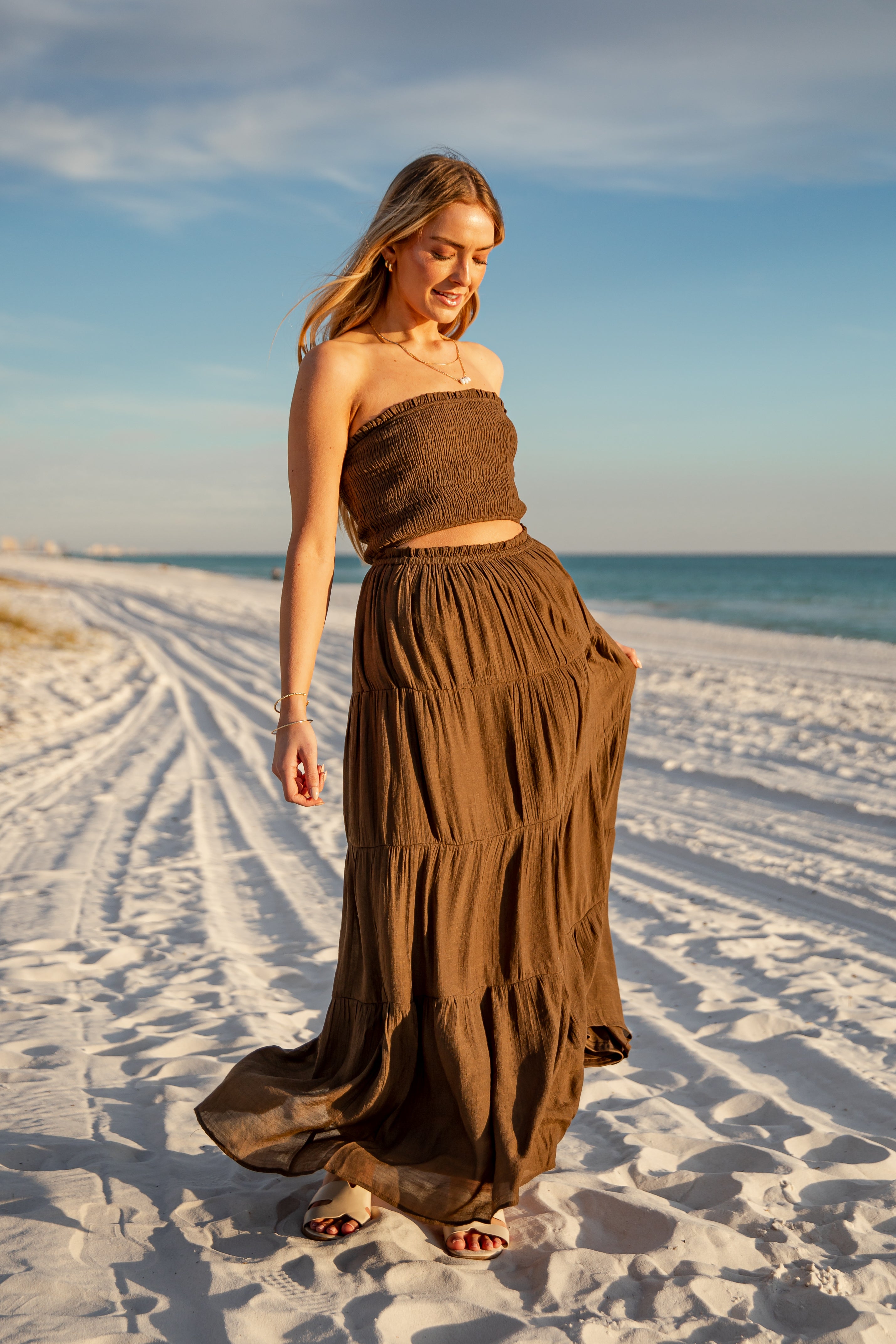Woman in a brown dress walking on a sandy beach with ocean and sky in the background