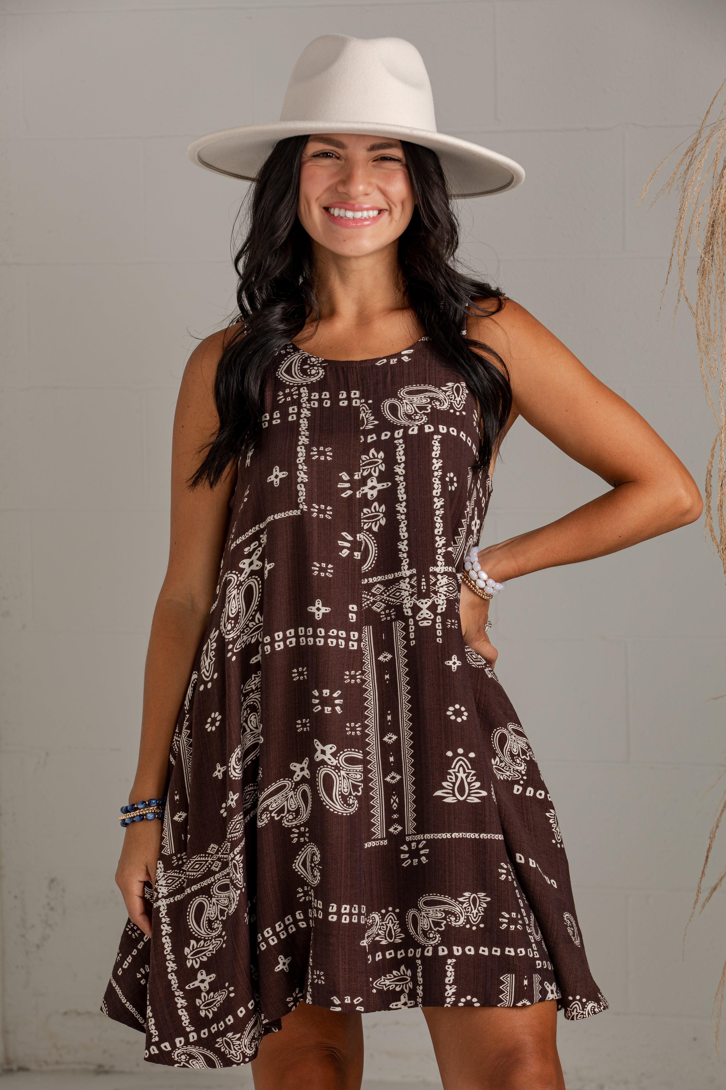 A woman wearing a sleeveless brown dress with a white paisley and geometric pattern, a wide-brimmed cream hat, and bracelets, posing against a light gray brick wall.