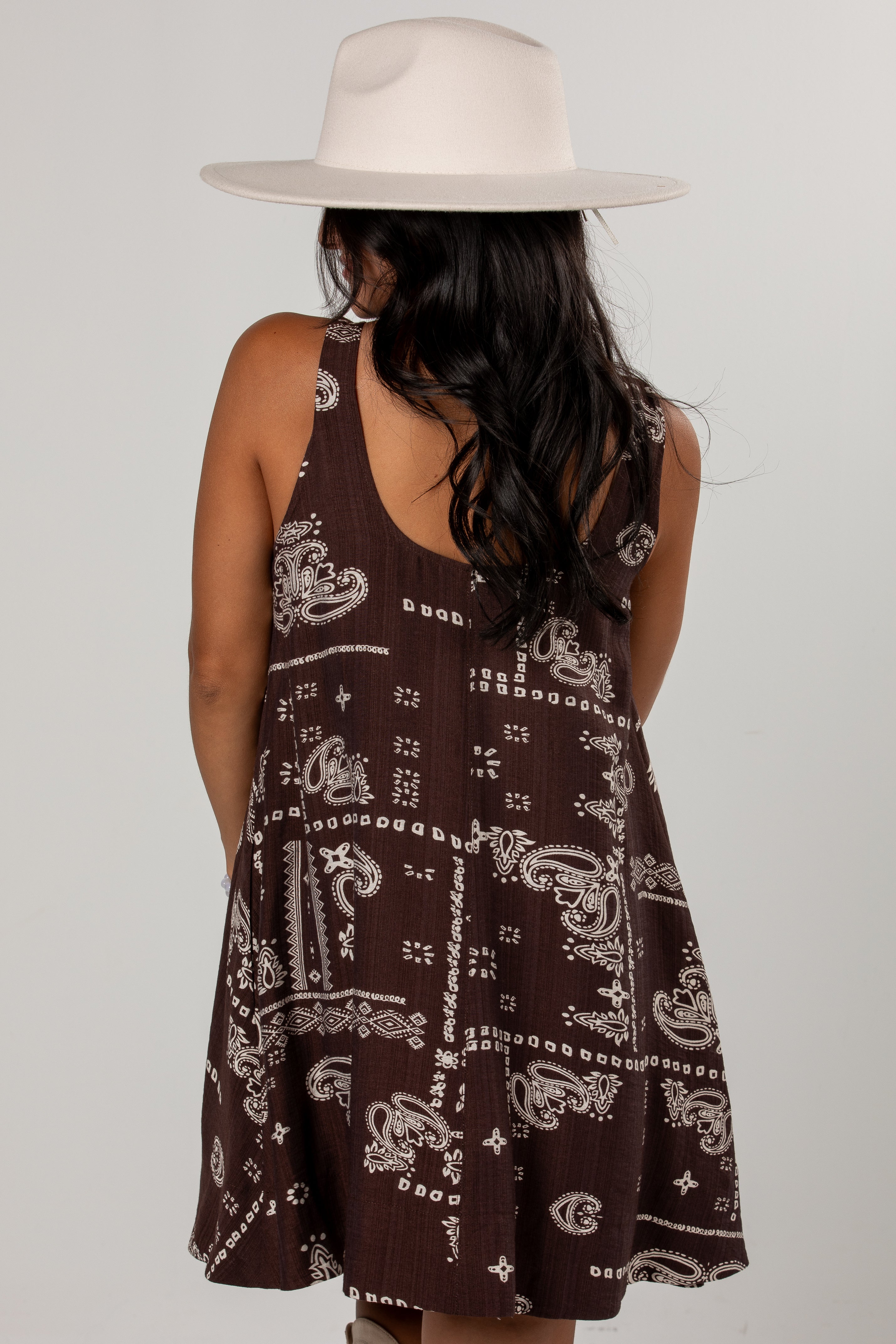 A woman viewed from behind wearing a sleeveless brown dress with a white paisley bandana-style print and a wide-brim cream hat, against a light gray studio background.
