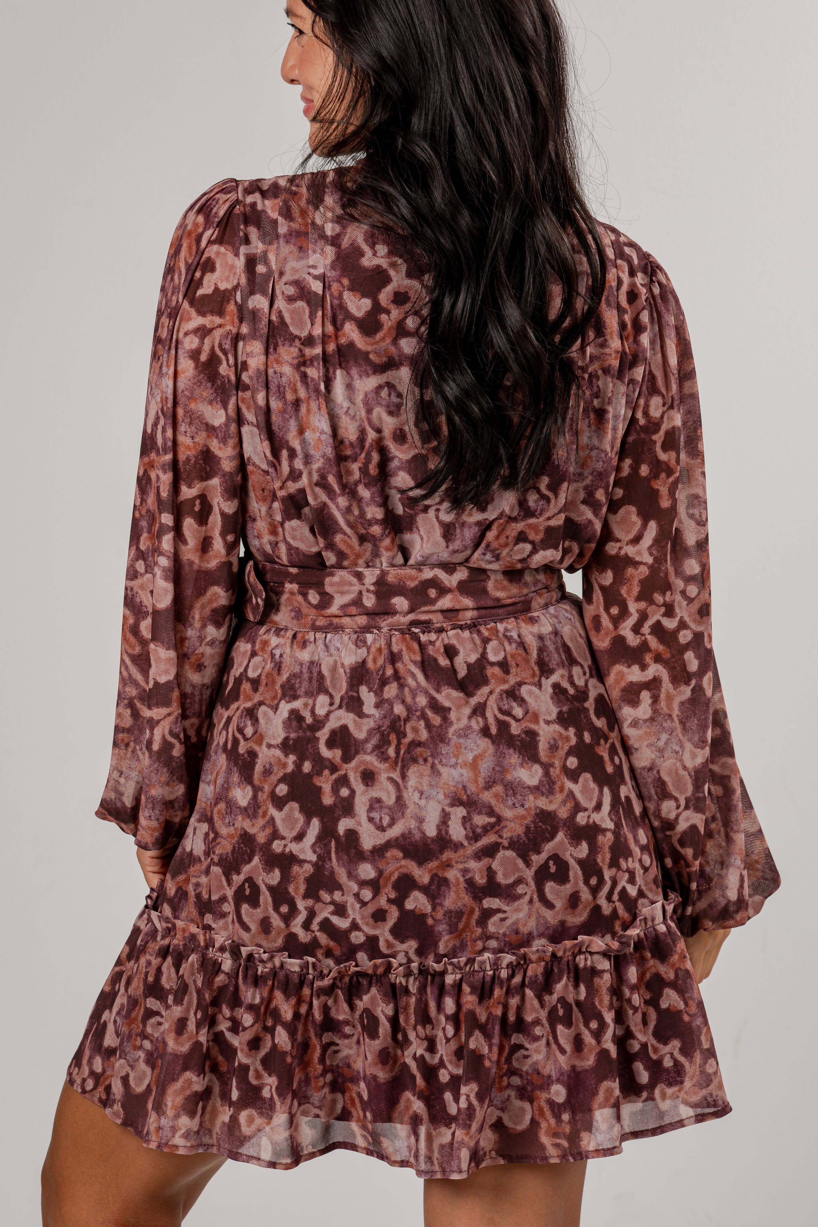 A woman shown from the back wearing a long-sleeve mini dress with a ruffled hem and waist tie, featuring an abstract floral pattern in dusty rose, mauve, and burgundy tones.