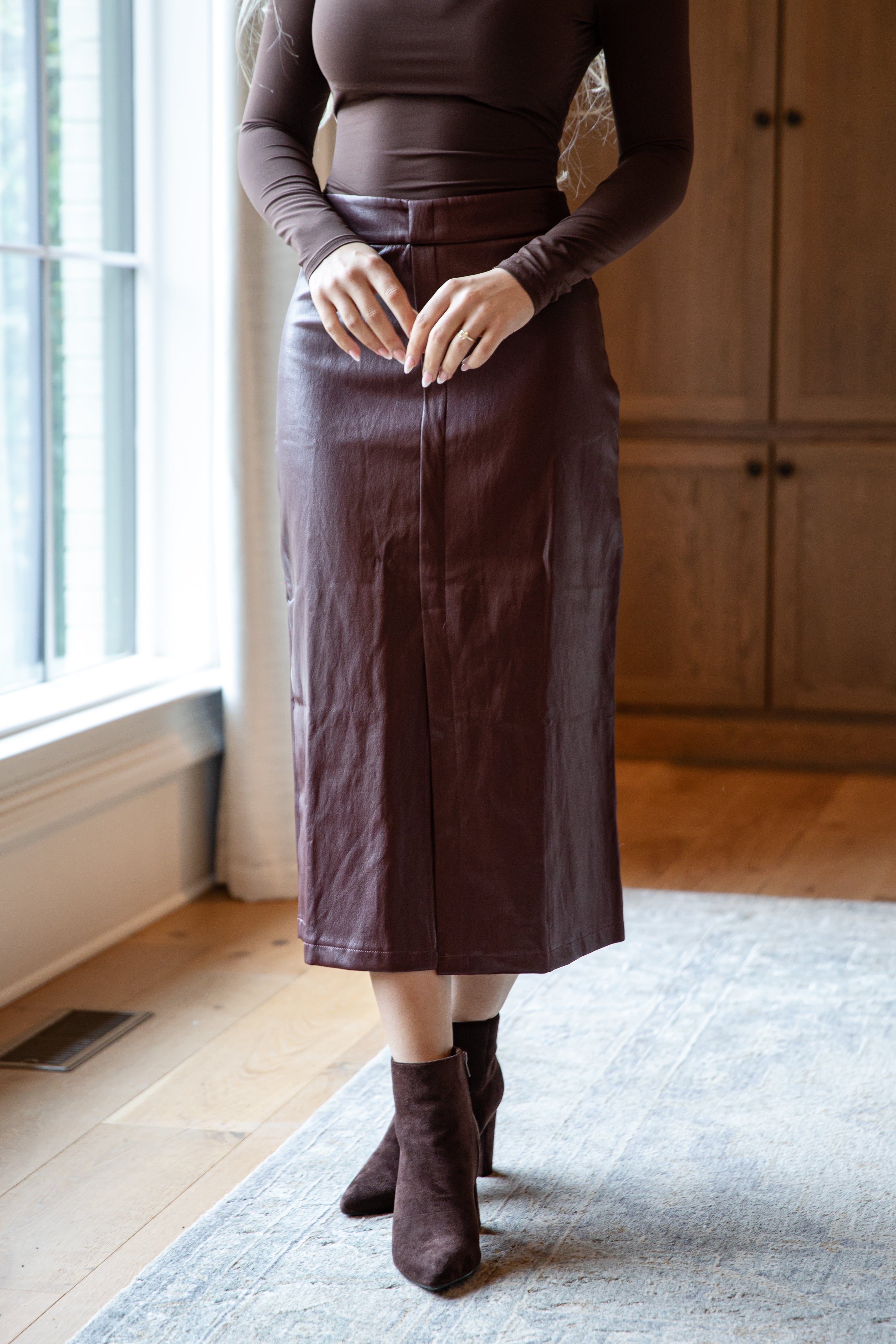 Person wearing a brown top and skirt standing in a room with wooden flooring and a window.