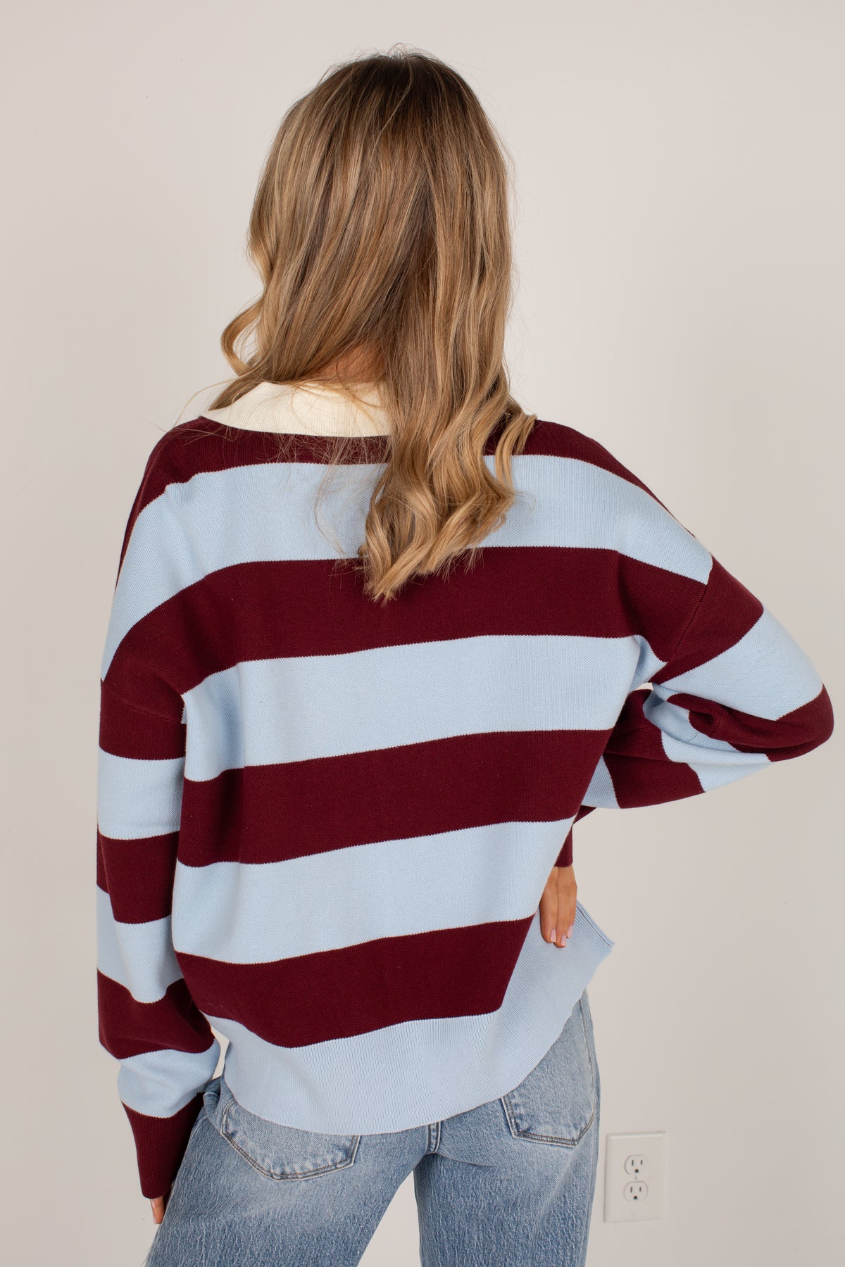 A woman viewed from behind wearing a long-sleeve knit sweater with wide horizontal stripes in burgundy and light blue, paired with light-wash jeans against a plain beige wall.