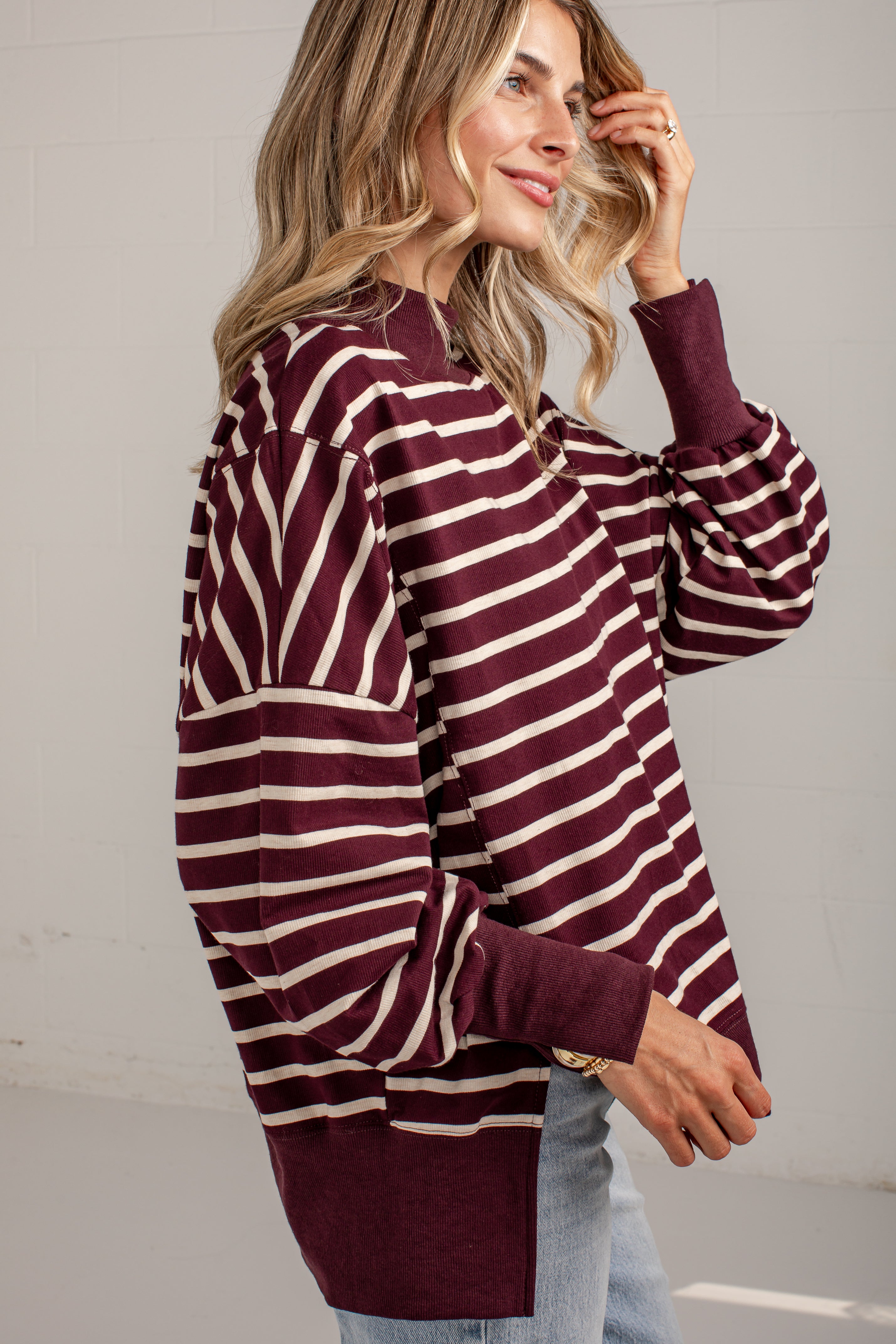 A woman wearing a long-sleeve knit sweater with horizontal burgundy and cream stripes, paired with light blue jeans, shown from a side profile against a light gray wall.