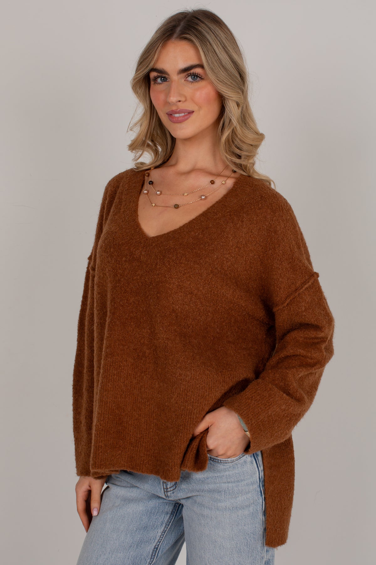 A woman wearing a fuzzy, oversized brown V-neck sweater and light blue jeans, standing against a solid light gray studio background.