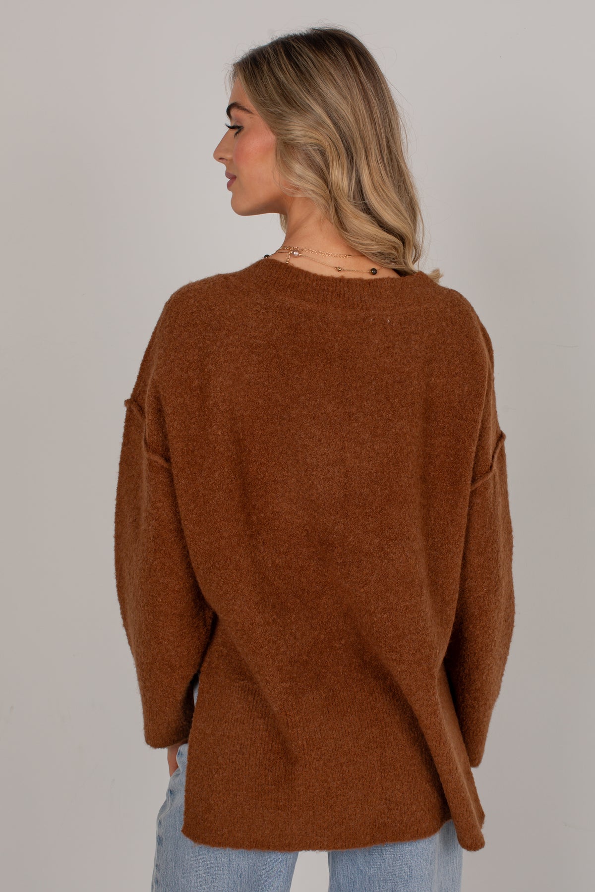 A woman shown from the back wearing a brown knit sweater and light blue jeans against a plain light gray studio background.
