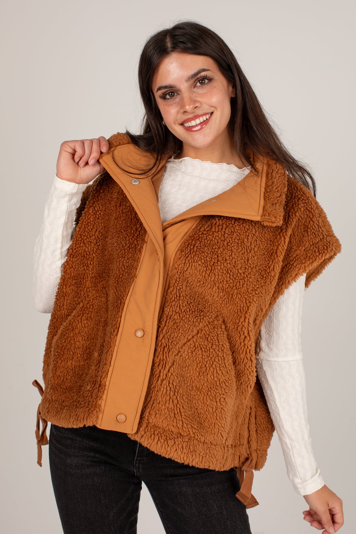 A smiling woman wearing a brown sherpa fleece vest over a white long-sleeve top and black jeans, standing against a light gray studio background.