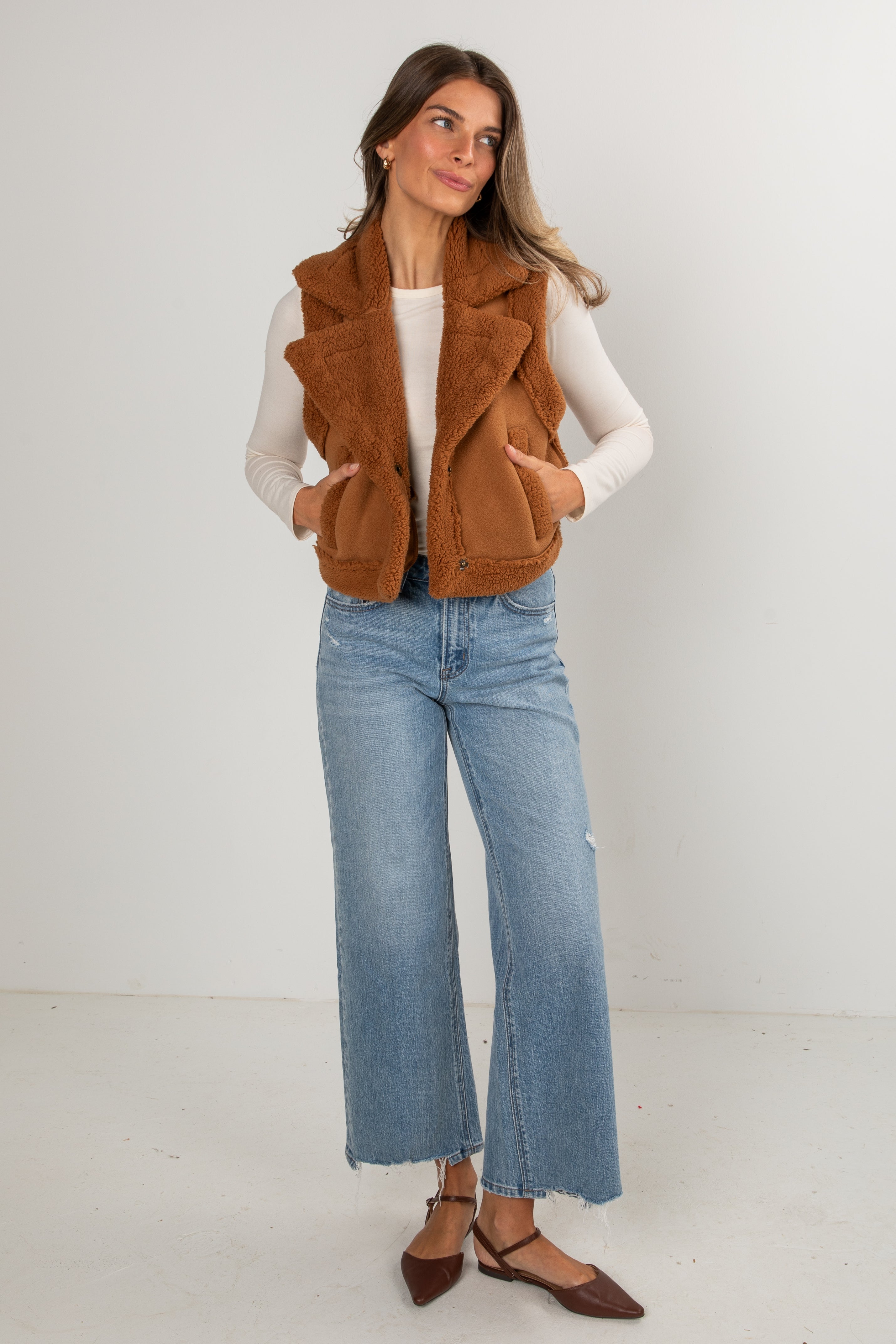 Woman wearing a brown vest, white shirt, and blue jeans against a white background