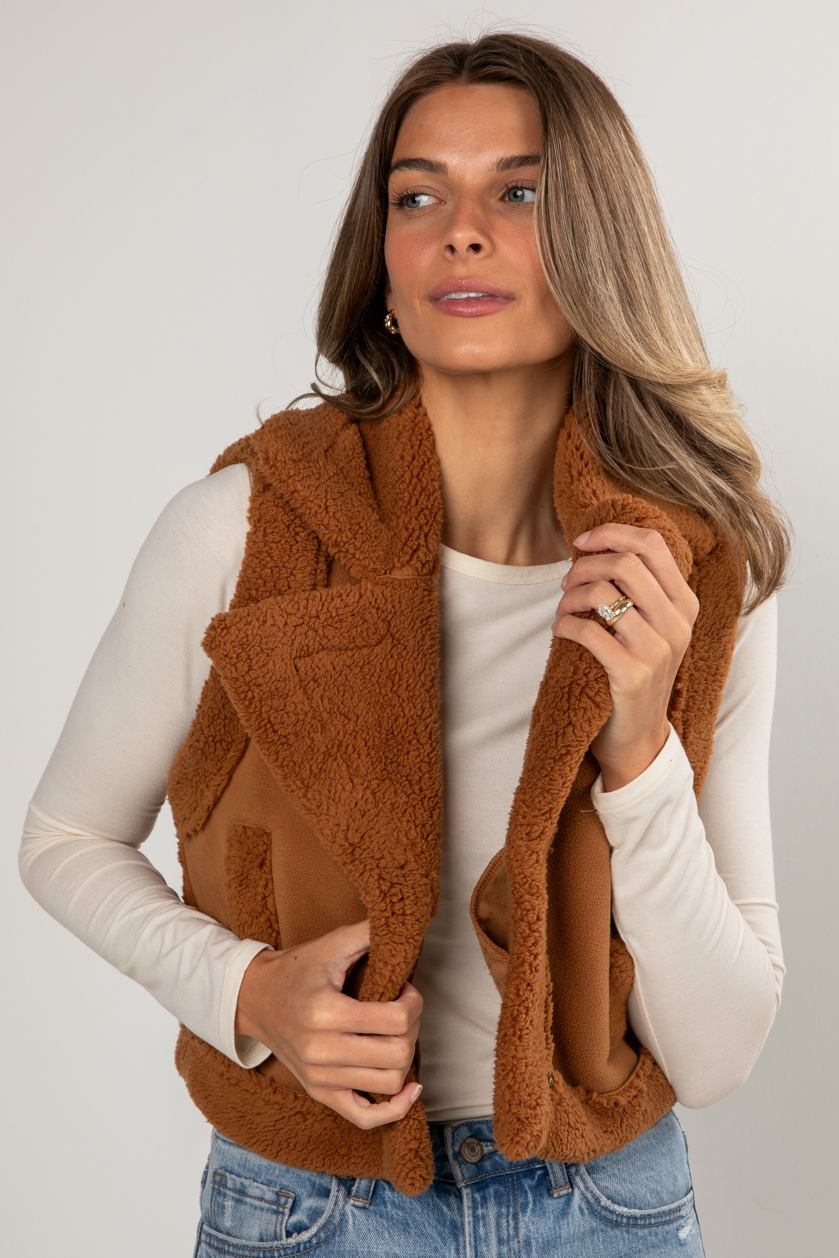 Woman wearing a brown fleece vest over a white shirt and jeans on a plain background