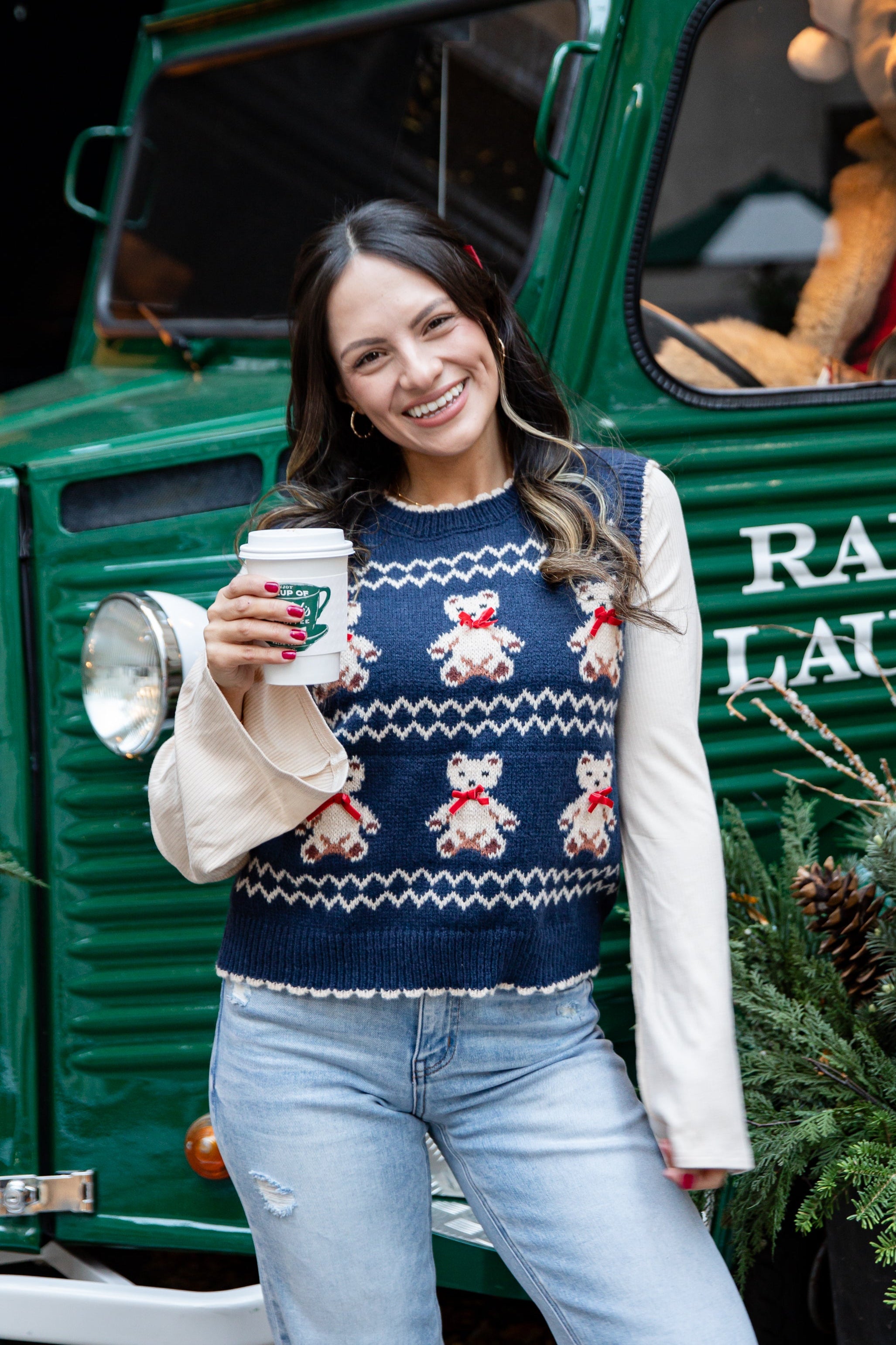 Woman in a festive sweater standing in front of a green truck with a teddy bear, holding a coffee cup.