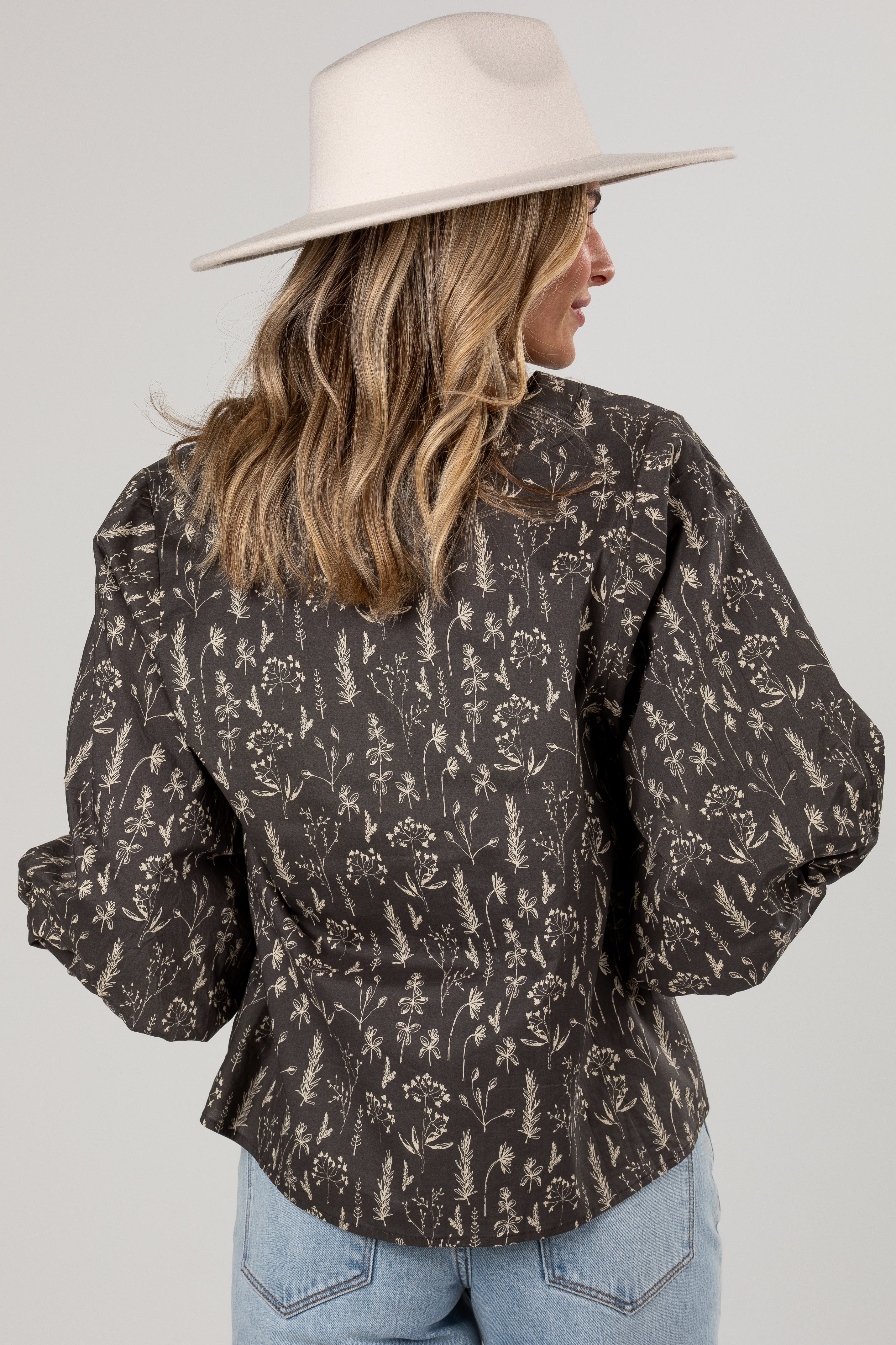 A woman shown from the back wearing a dark brown long-sleeve blouse with a beige botanical pattern, paired with light blue jeans and a cream wide-brim hat, against a light gray studio background.