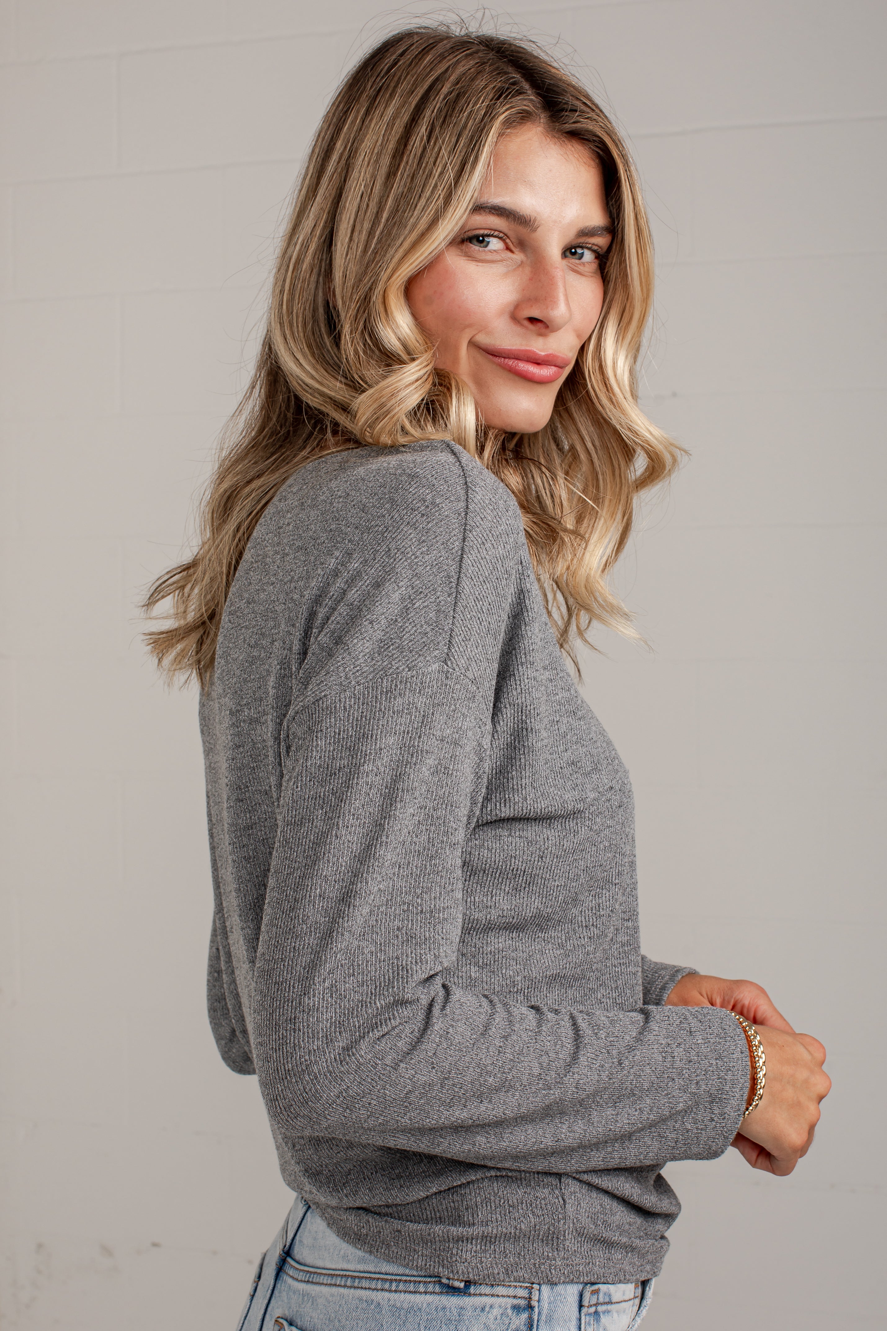 A woman with blonde wavy hair wearing a heather gray long-sleeve knit top and light blue jeans, photographed against a light gray wall.