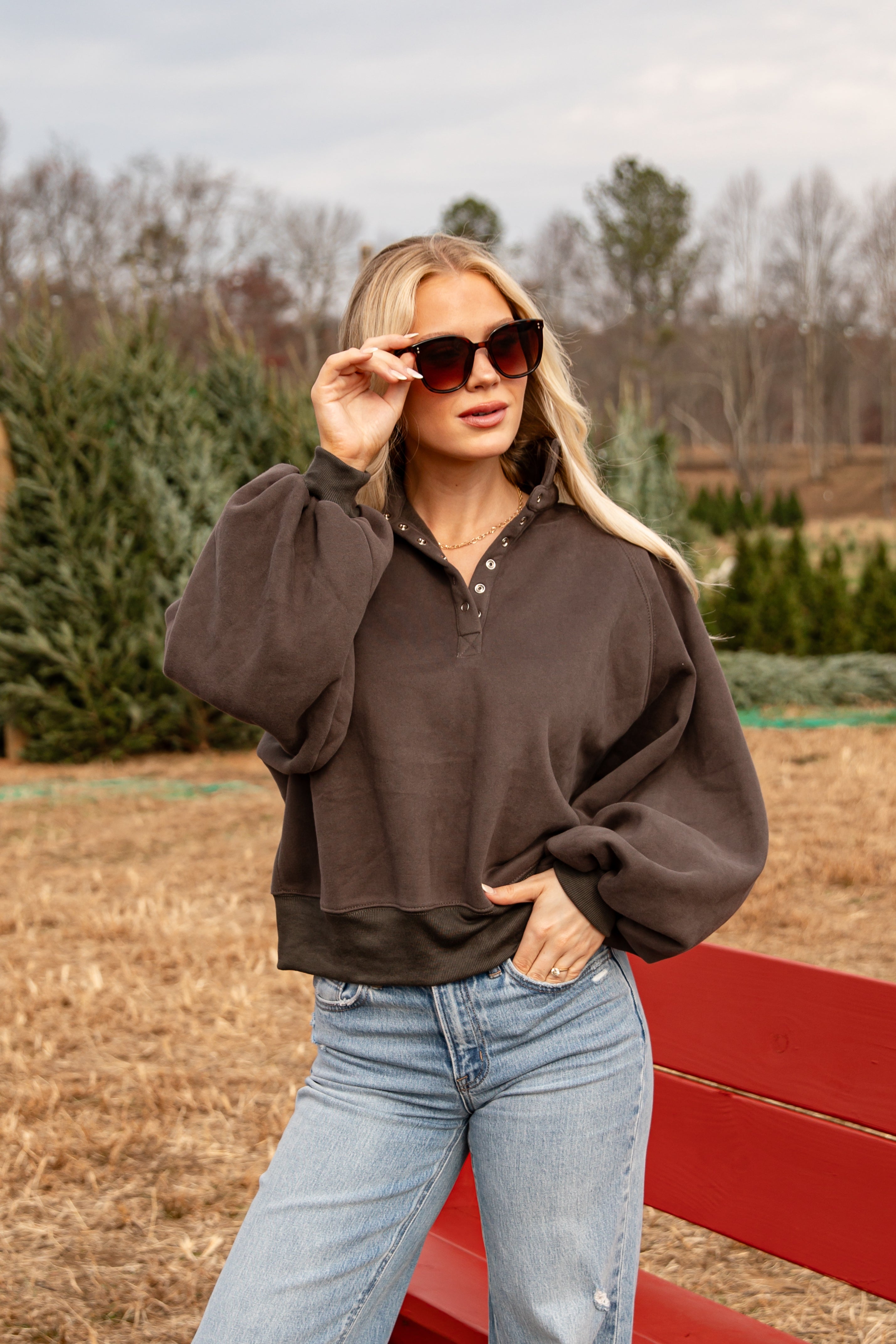 Woman wearing a brown sweater and blue jeans standing in a field with trees in the background