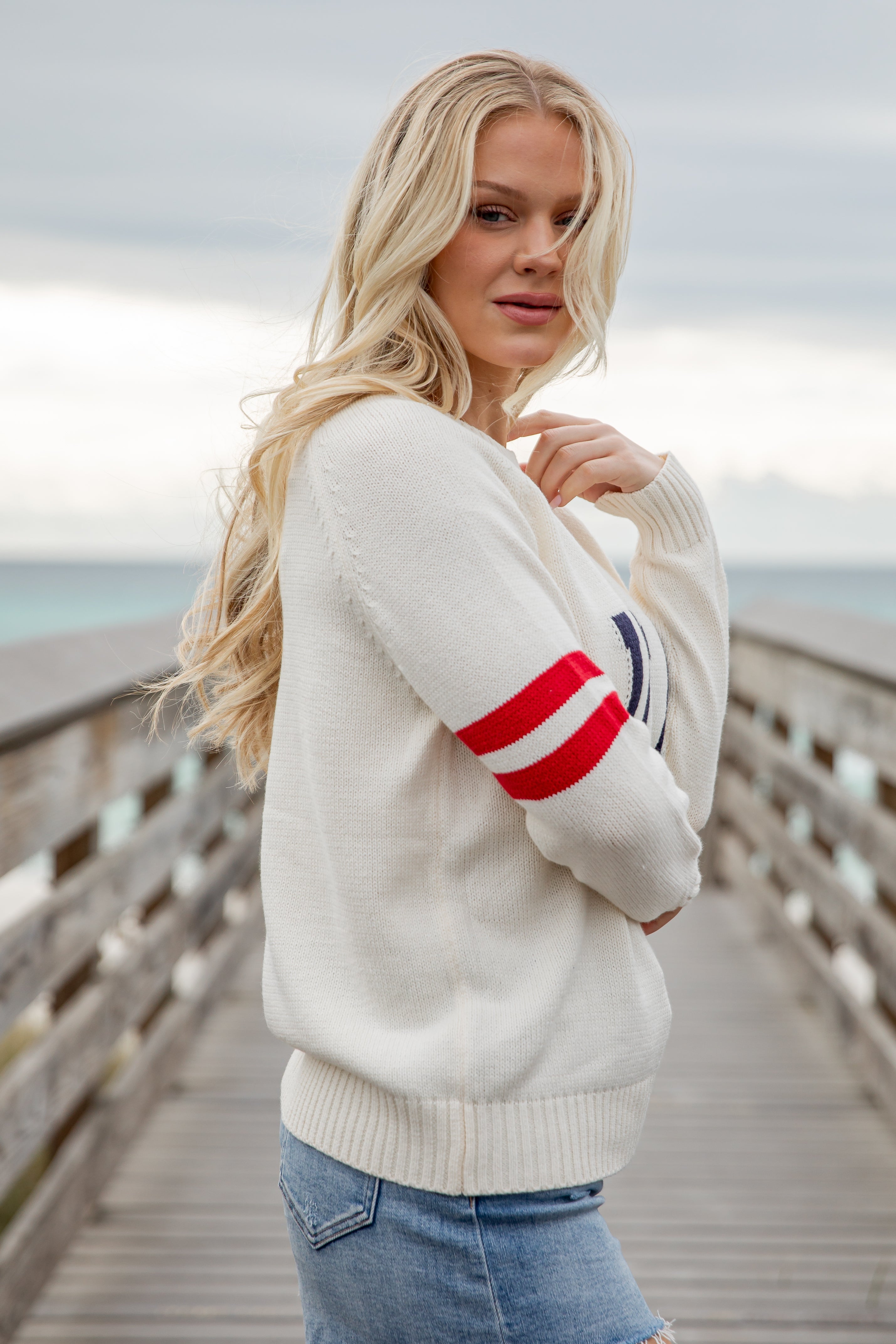 Woman wearing a white sweater with red and blue stripes on a wooden boardwalk by the sea.
