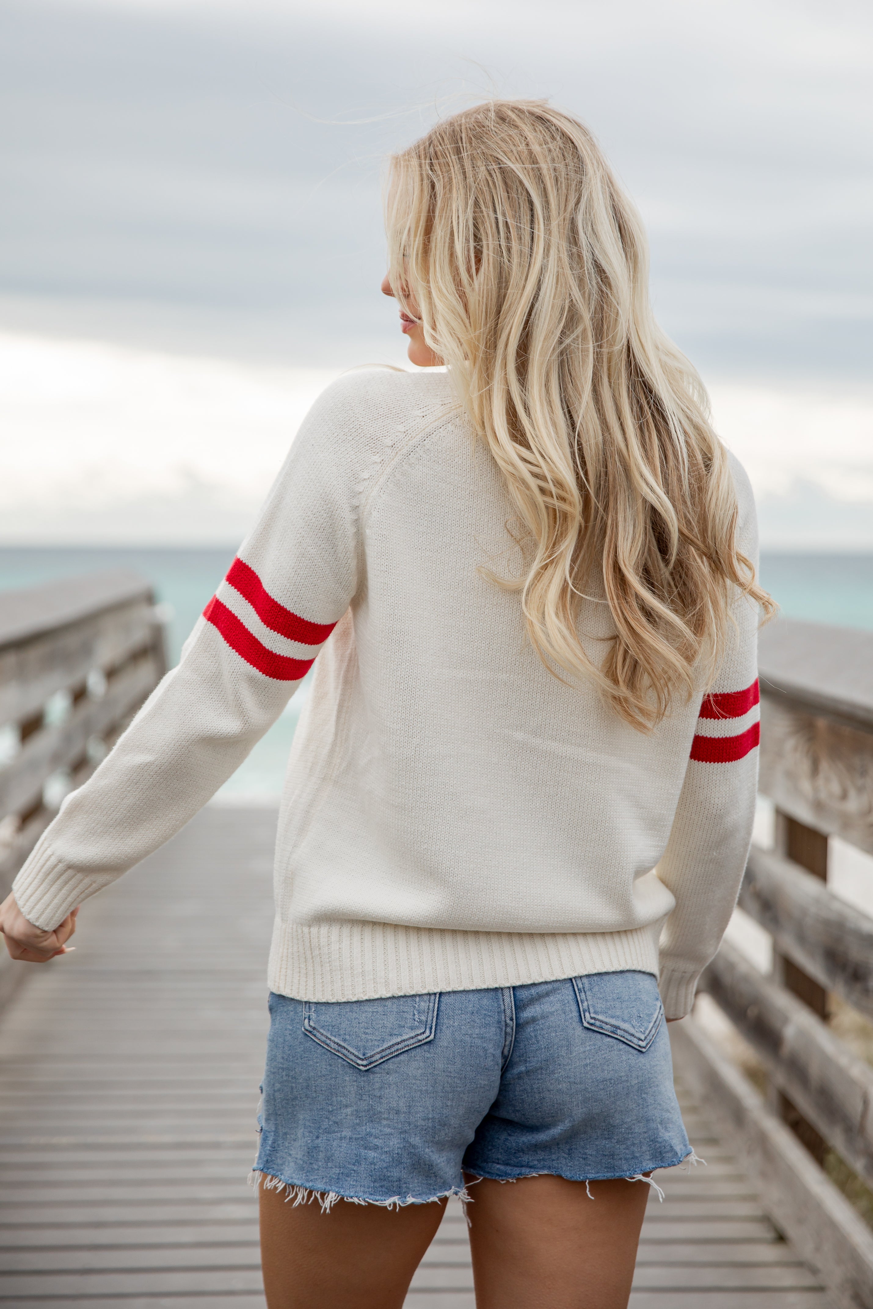 Woman wearing a cream sweater with red stripes and denim shorts, standing on a wooden boardwalk.