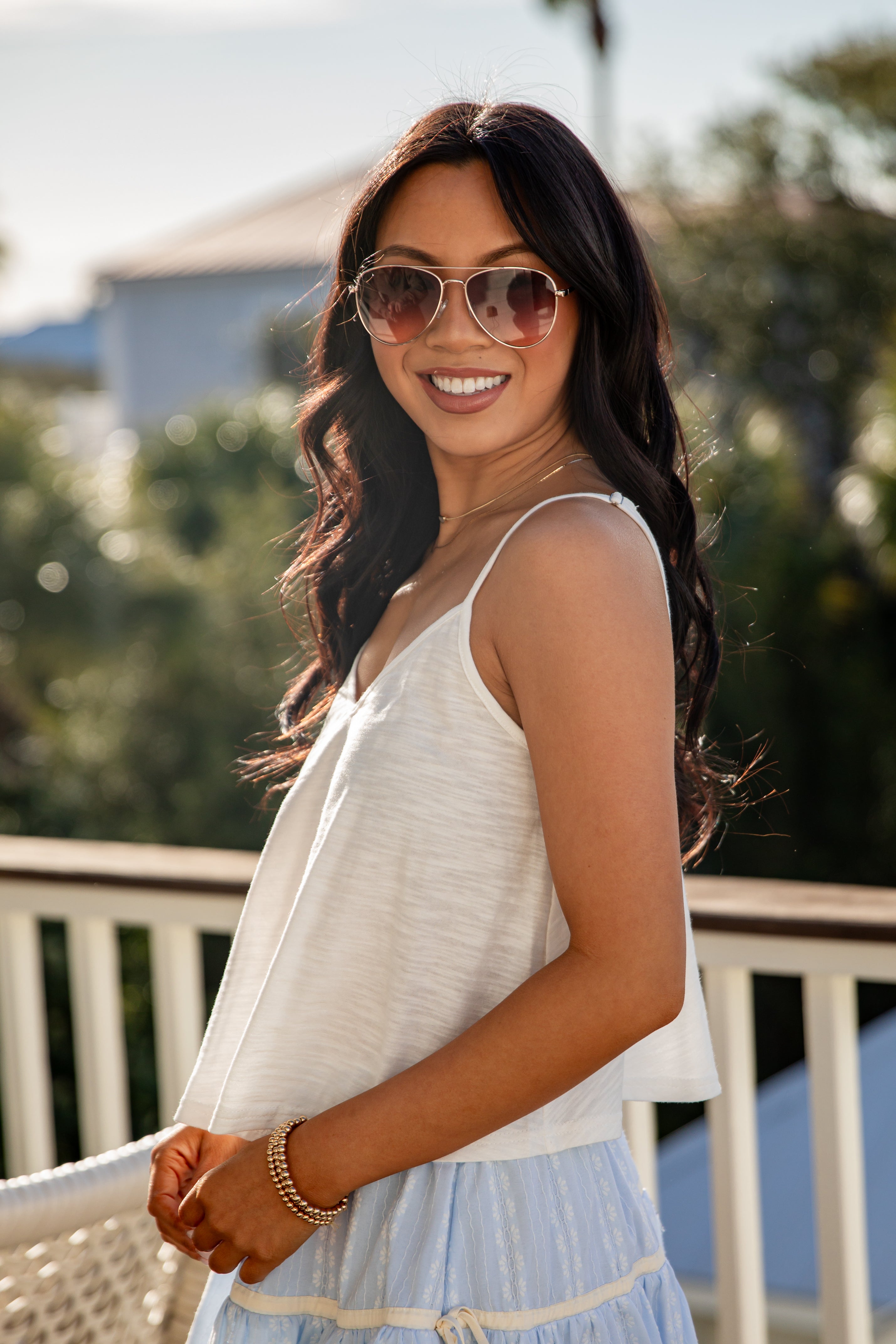 Woman wearing sunglasses and a white sleeveless top with a blurred outdoor background