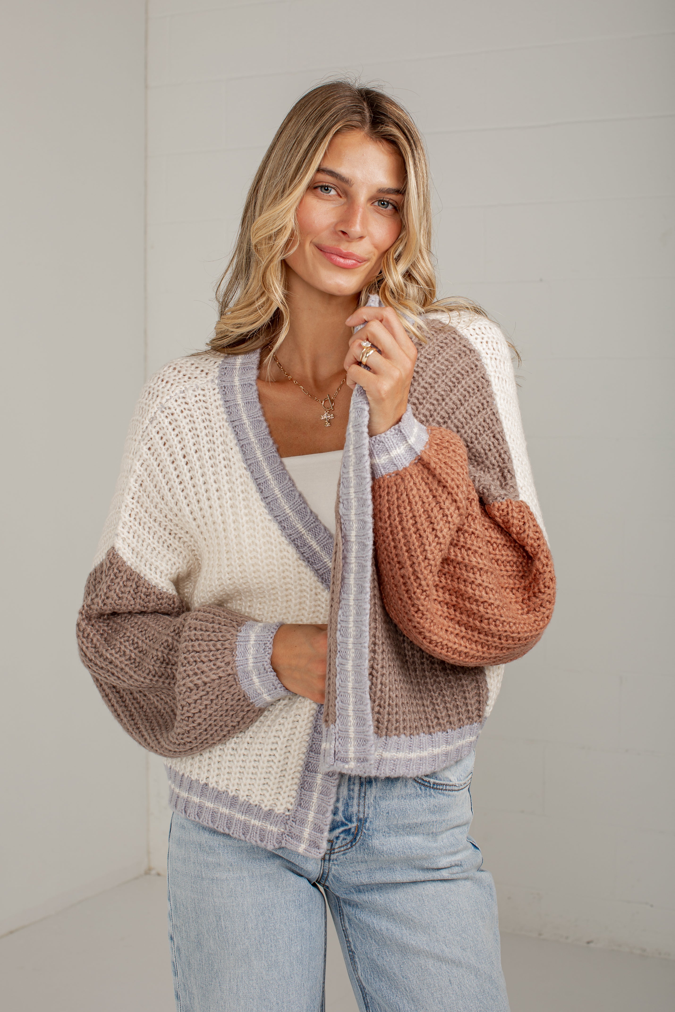 A woman wearing a chunky knit color-block cardigan with cream, taupe, and terracotta sections, paired with light-wash blue jeans, standing against a light gray wall.