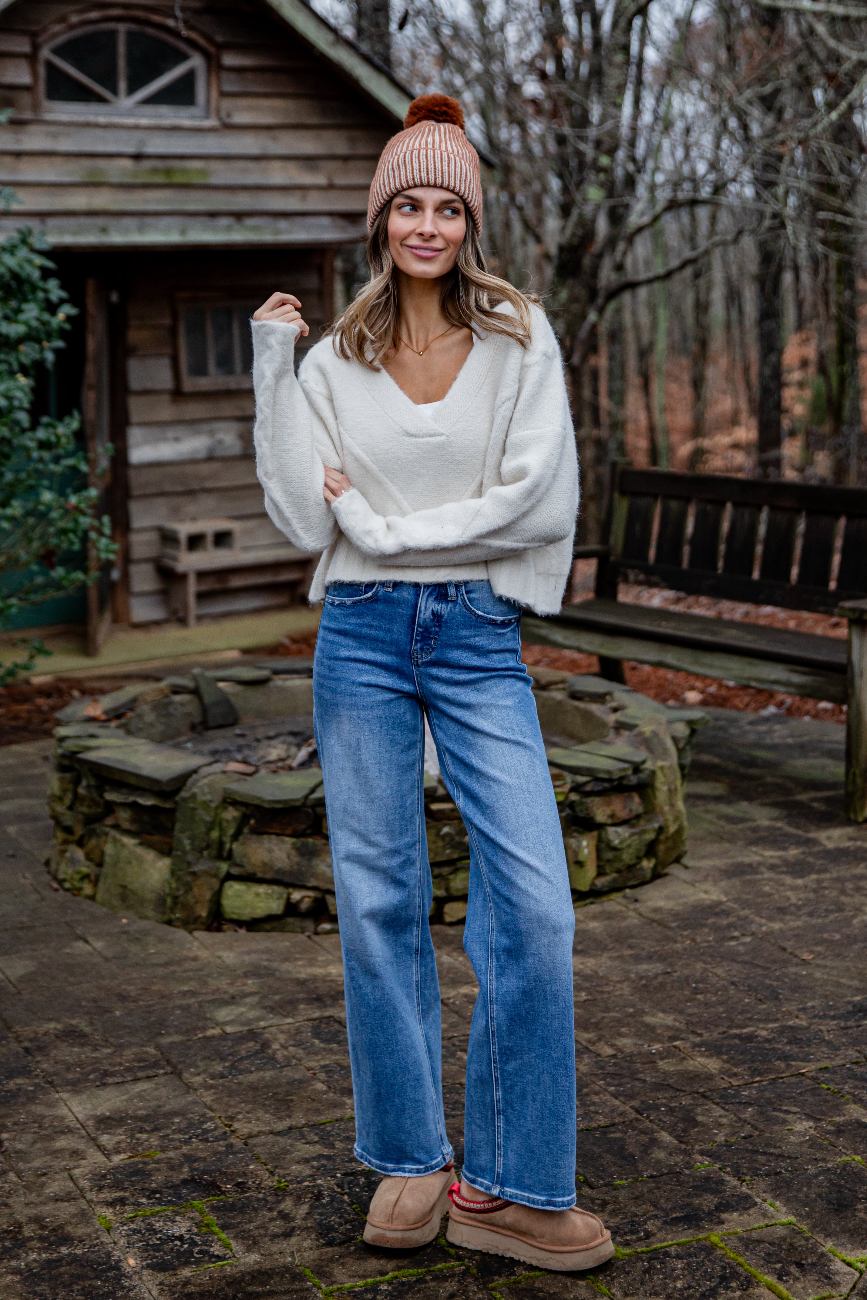 Woman wearing a white sweater, blue jeans, and a pink beanie standing in front of a wooden cabin.
