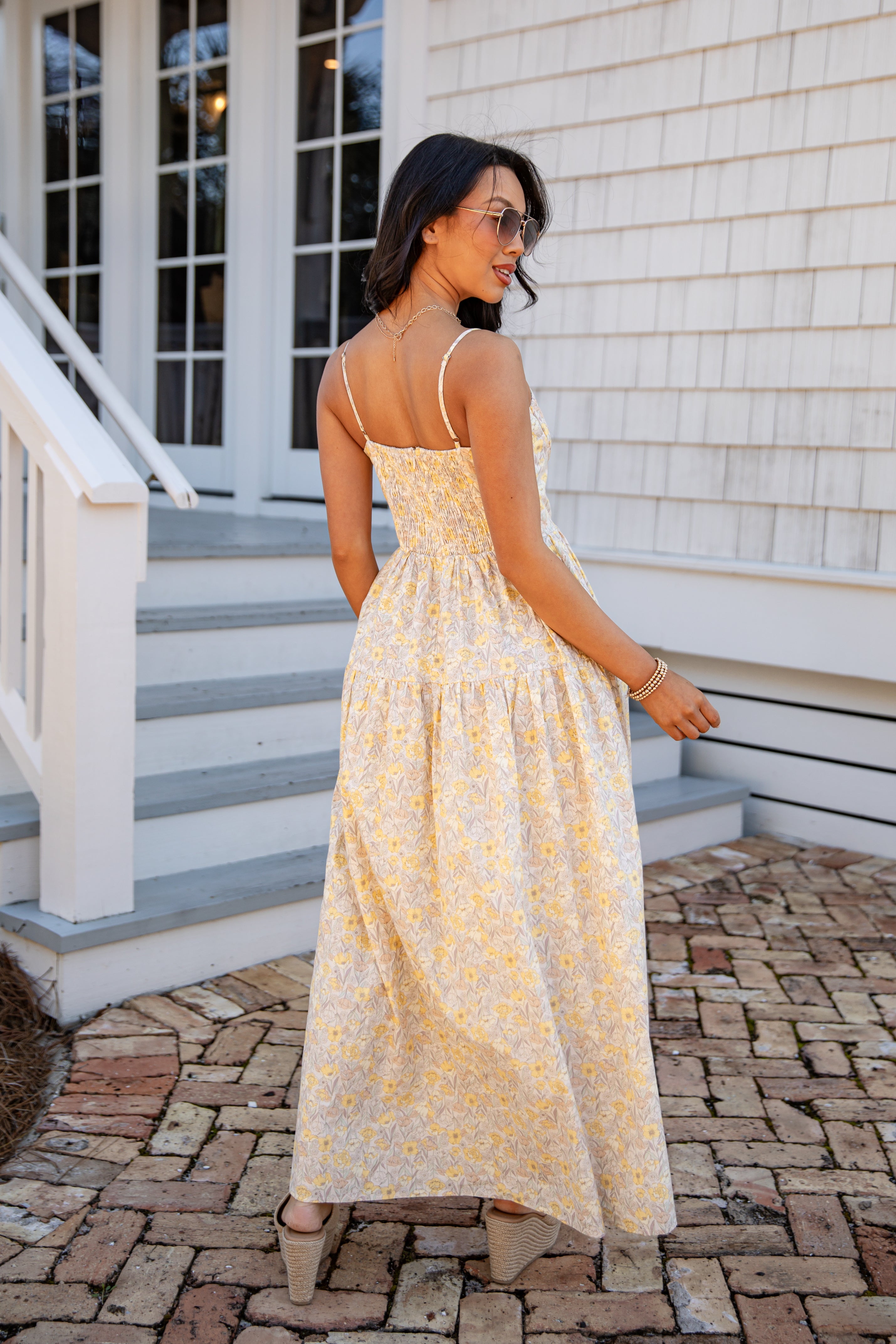 Woman in a yellow floral dress standing on a stone path in front of a house.