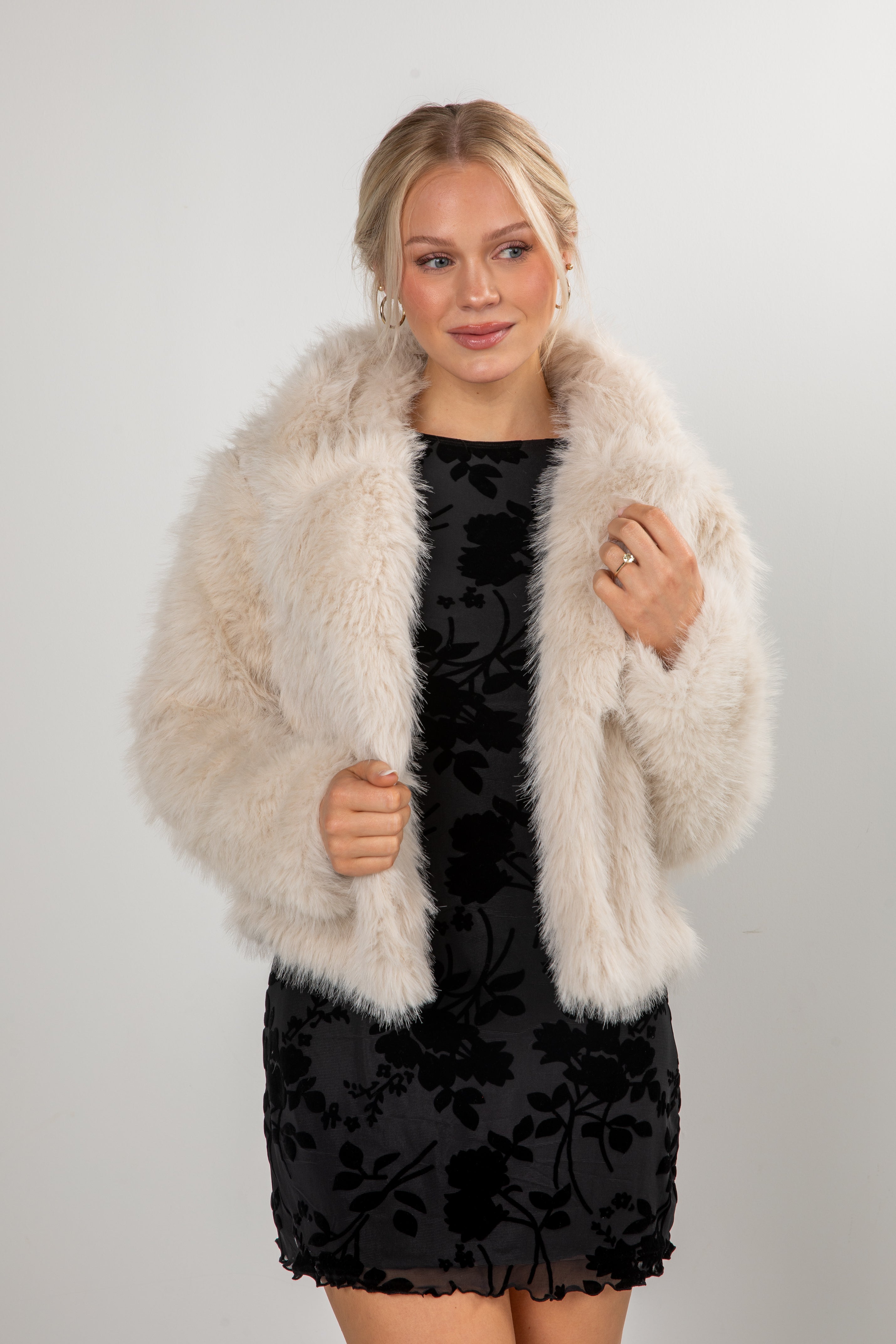 Woman wearing a black dress with floral patterns and a beige fur coat against a white background