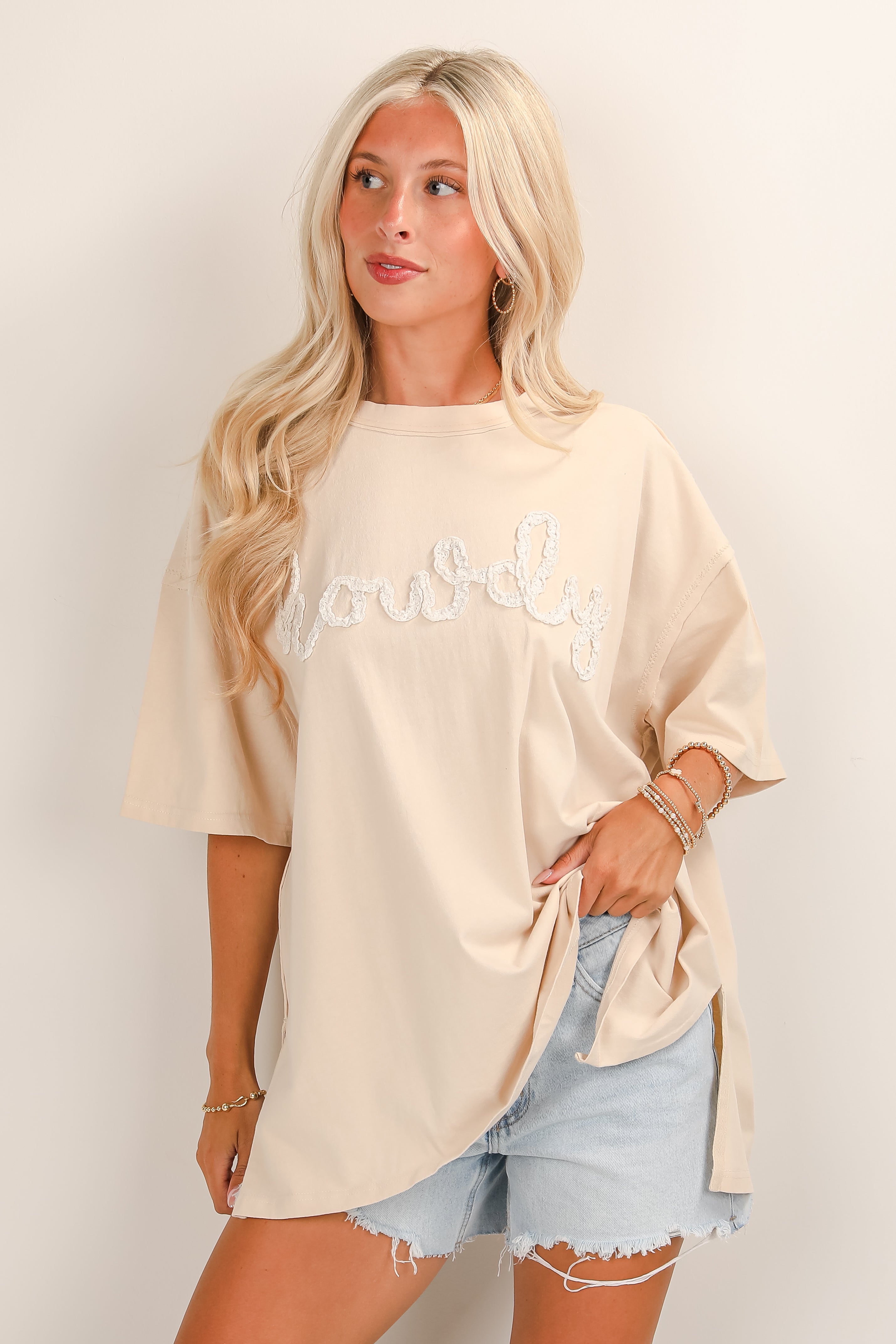 Woman wearing a beige oversized t-shirt with text and denim shorts on a plain background
