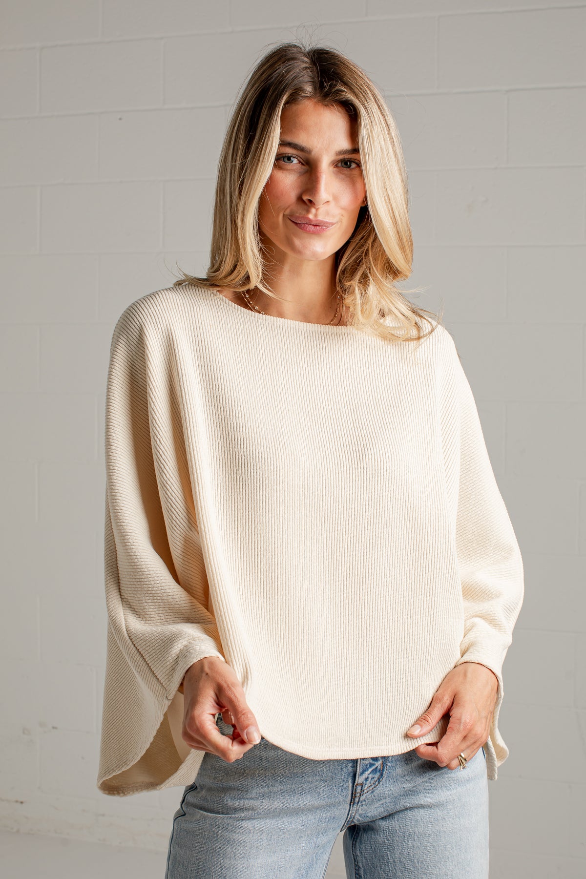 A woman wearing a cream-colored, long-sleeved ribbed knit top with a wide boat neckline and draped sleeves, paired with light-wash blue jeans, standing against a white brick wall.