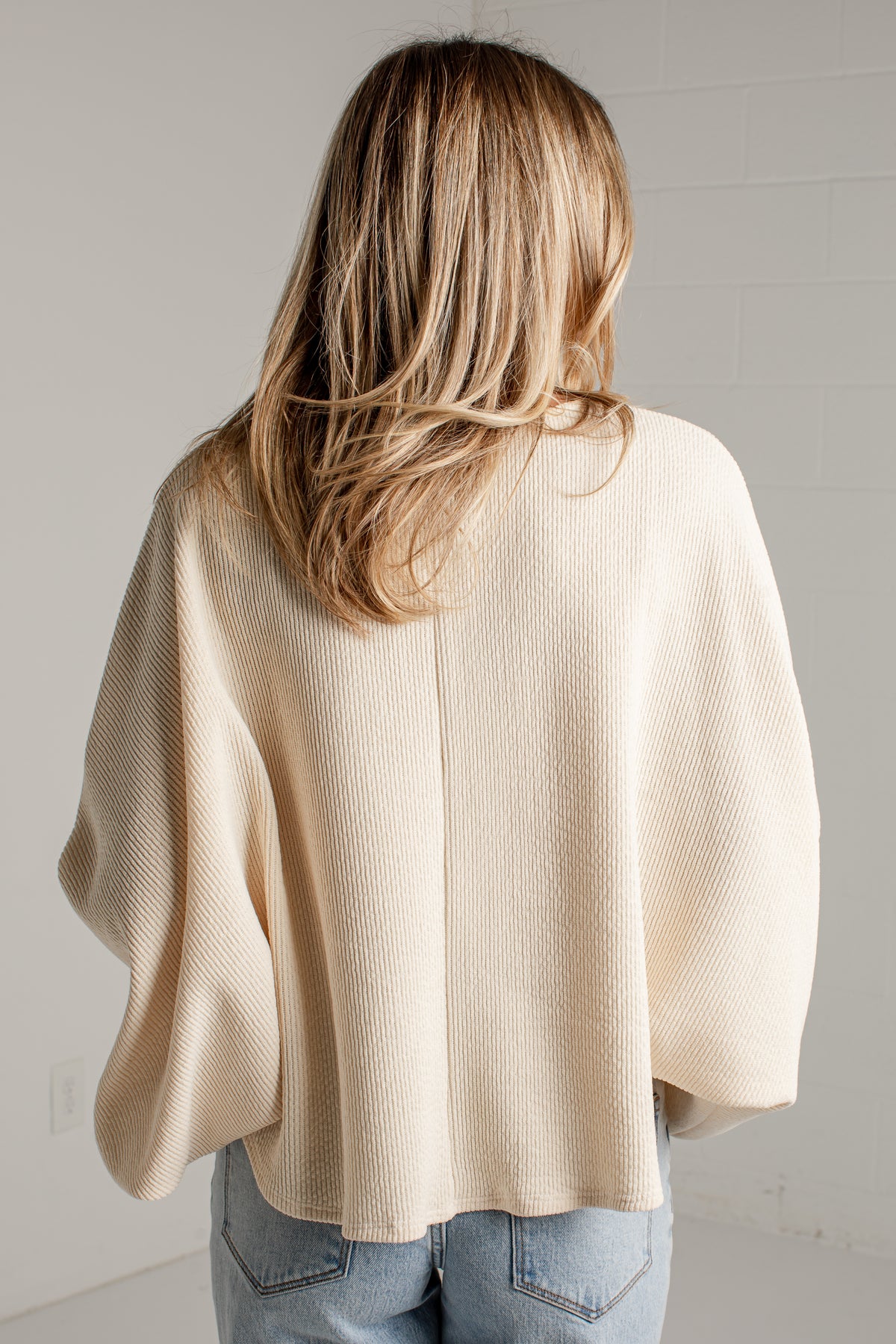 A woman viewed from behind wearing a cream-colored, textured knit poncho sweater and light blue jeans against a white brick wall.