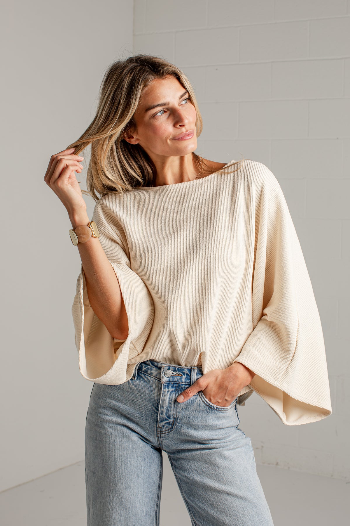 A woman wearing a cream-colored, wide-sleeved bubble-sleeve top and light-wash blue jeans, standing against a white brick wall.