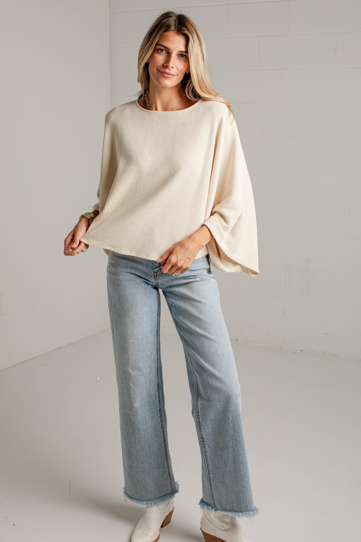 A woman modeling a cream-colored, oversized long-sleeve knit top with wide bell sleeves, paired with light-wash blue jeans and white boots, standing in a bright studio setting.