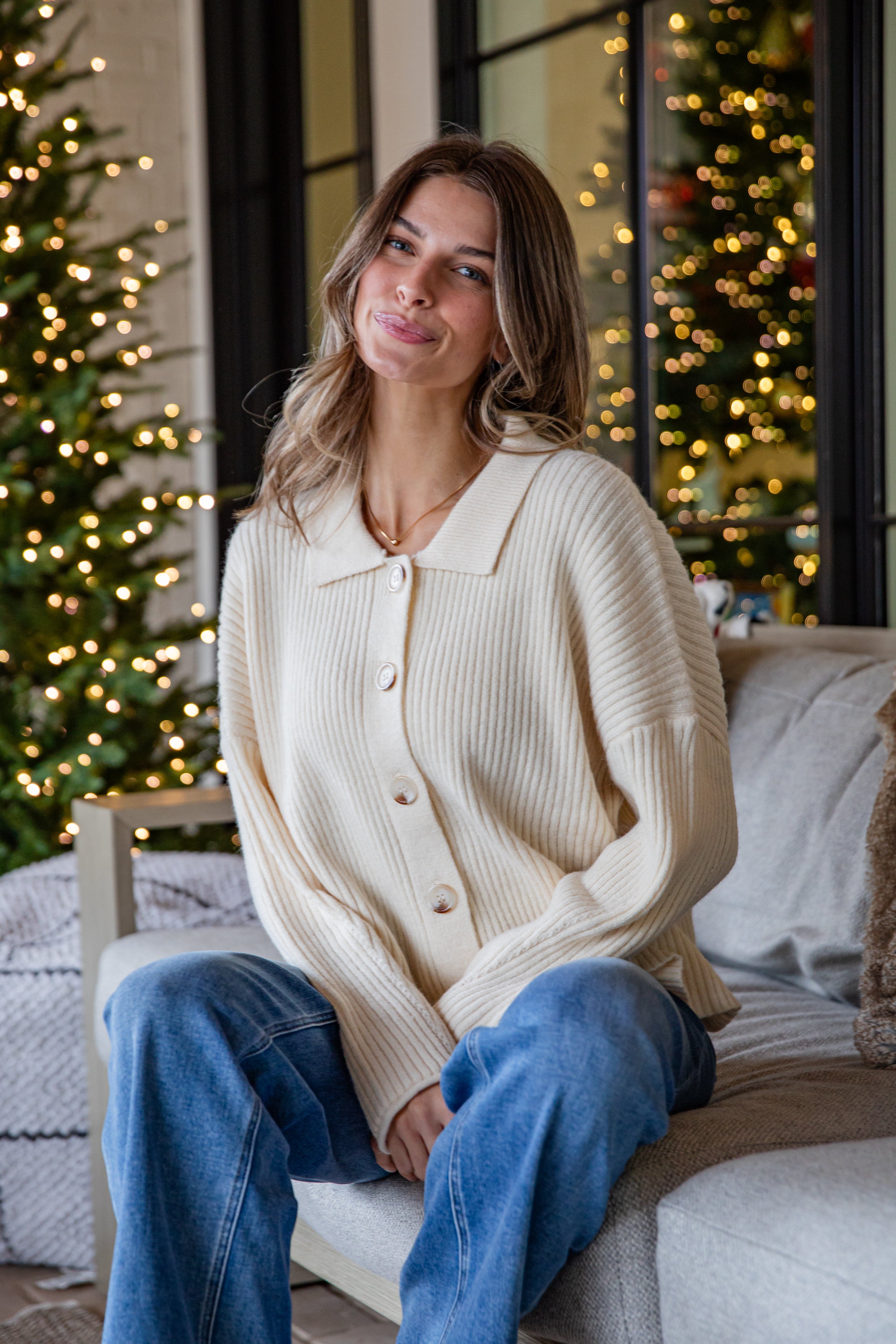 Woman wearing a cream cardigan and blue jeans sitting on a couch with Christmas trees in the background