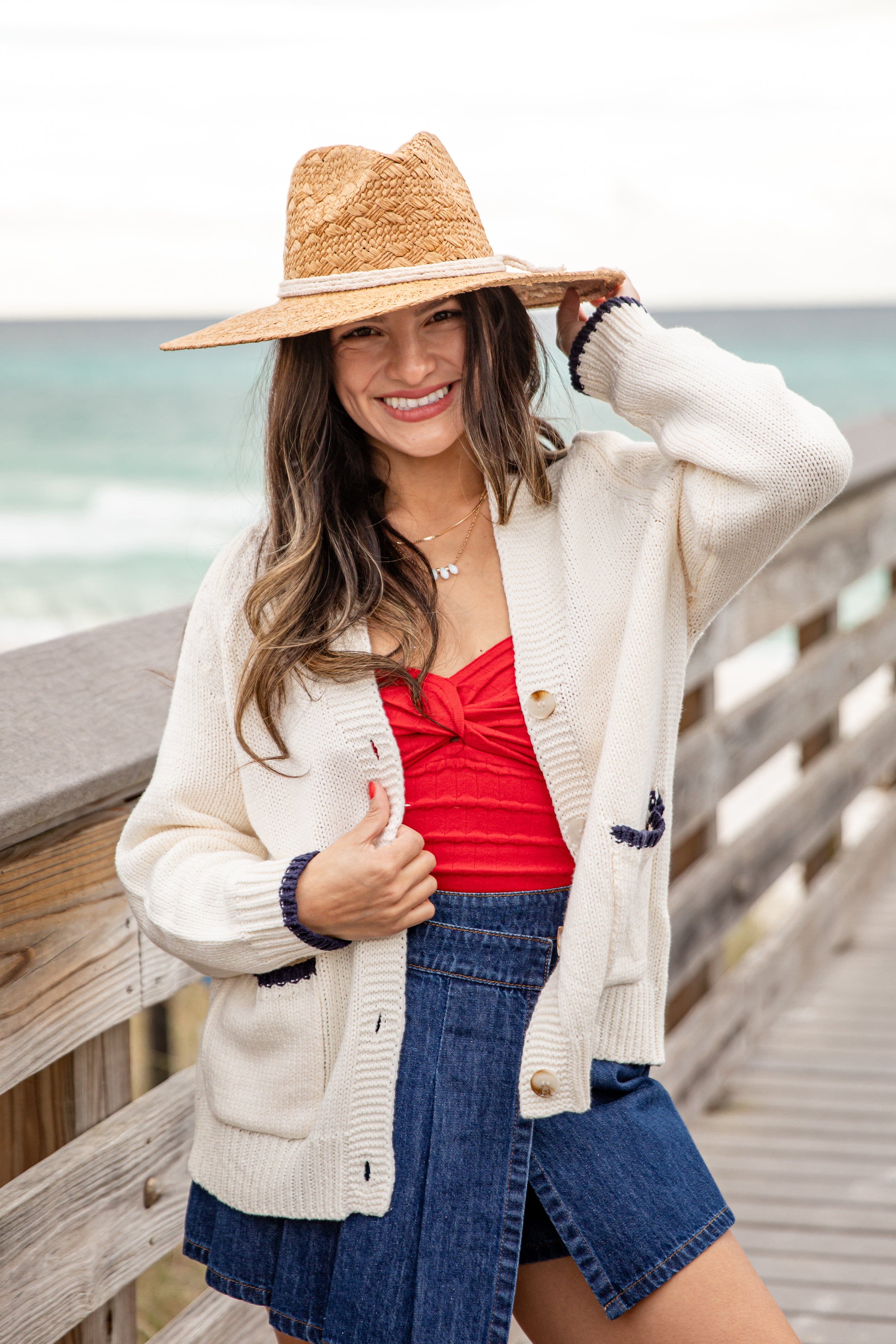 Woman wearing a straw hat, white cardigan, red top, and denim shorts by the beach.