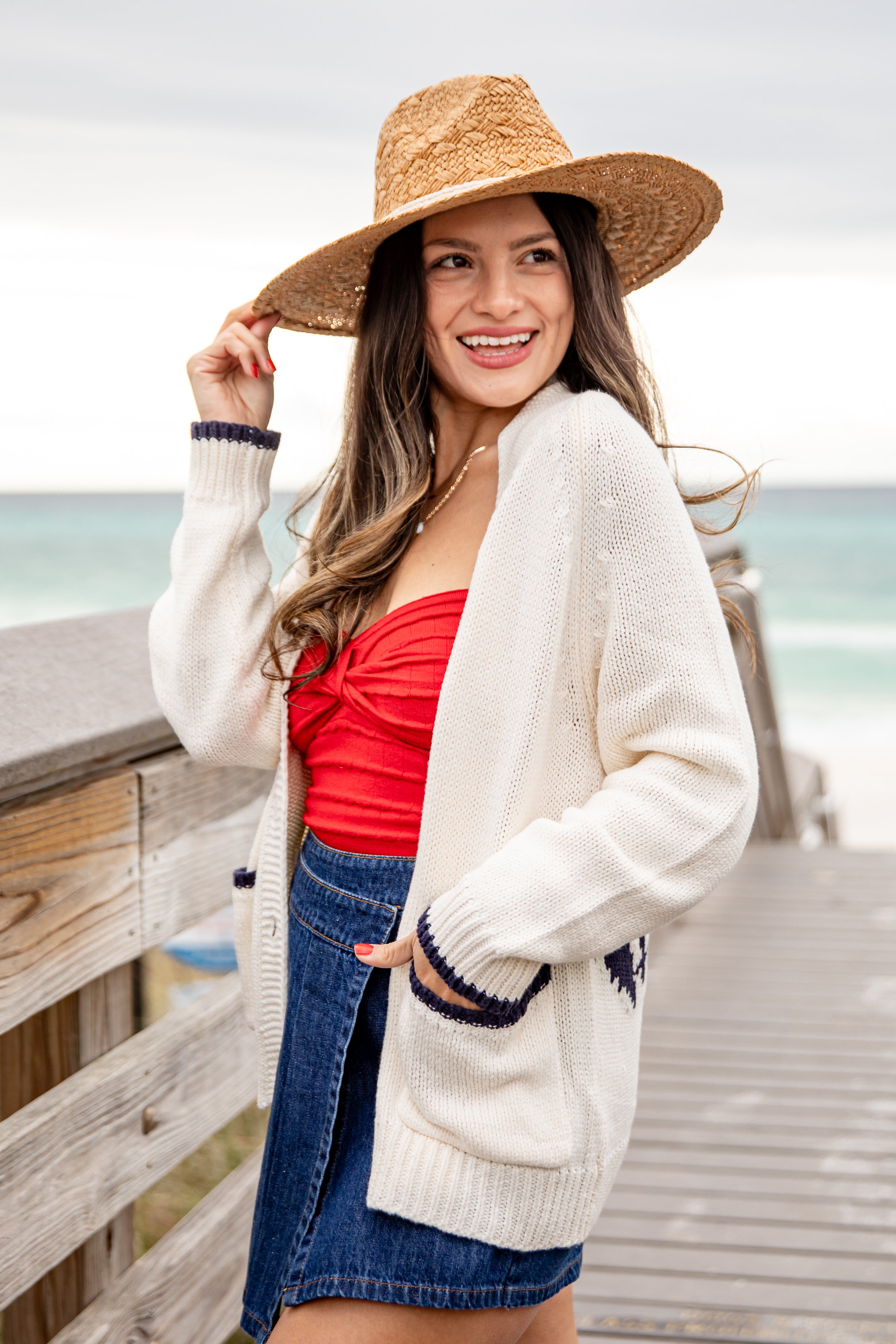 Woman wearing a straw hat, white cardigan, red top, and blue skirt on a wooden deck by the ocean.