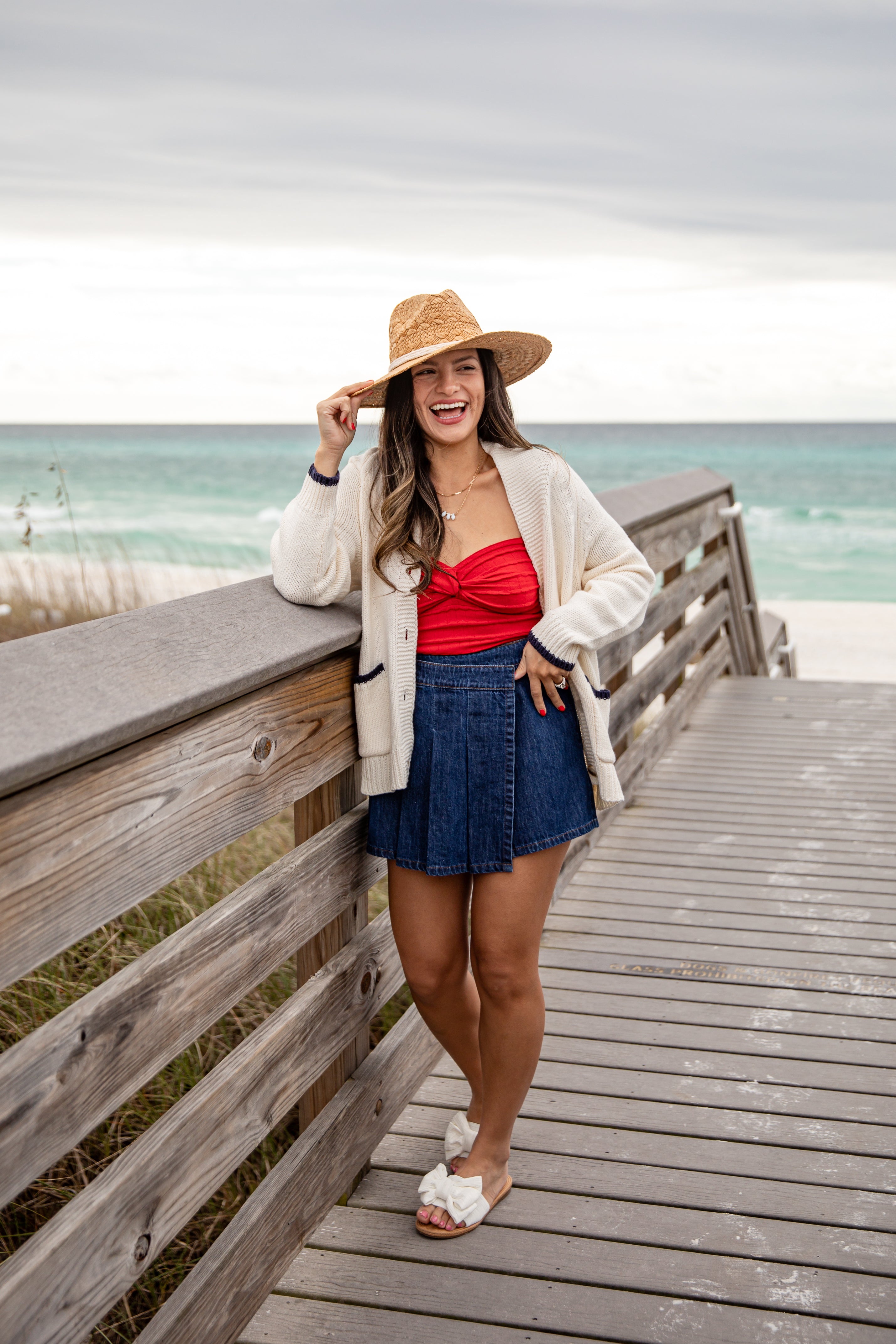 Woman standing on a wooden boardwalk by the ocean, wearing a straw hat, red top, and denim skirt.