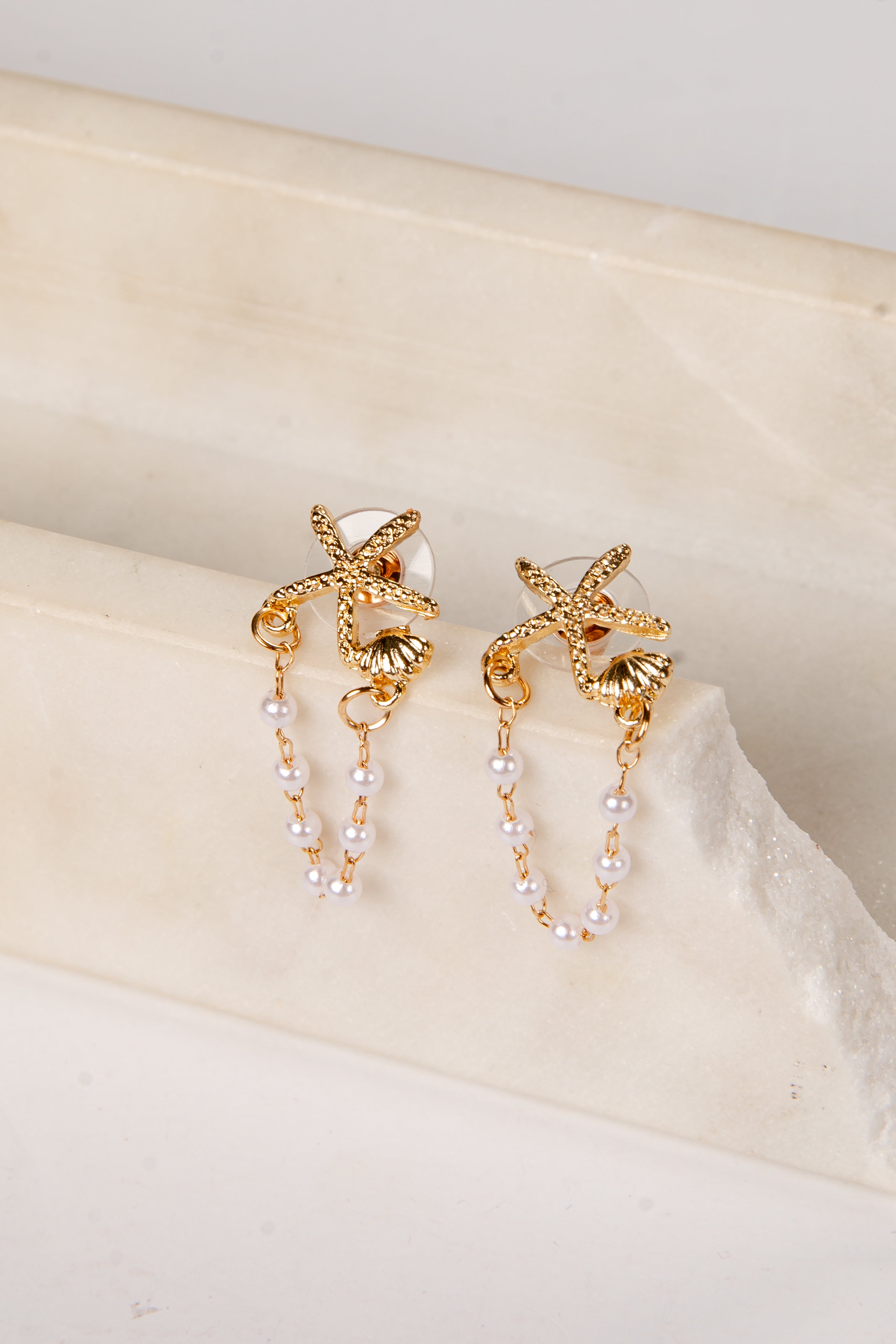 Gold earrings with starfish designs and pearl strands on a beige surface